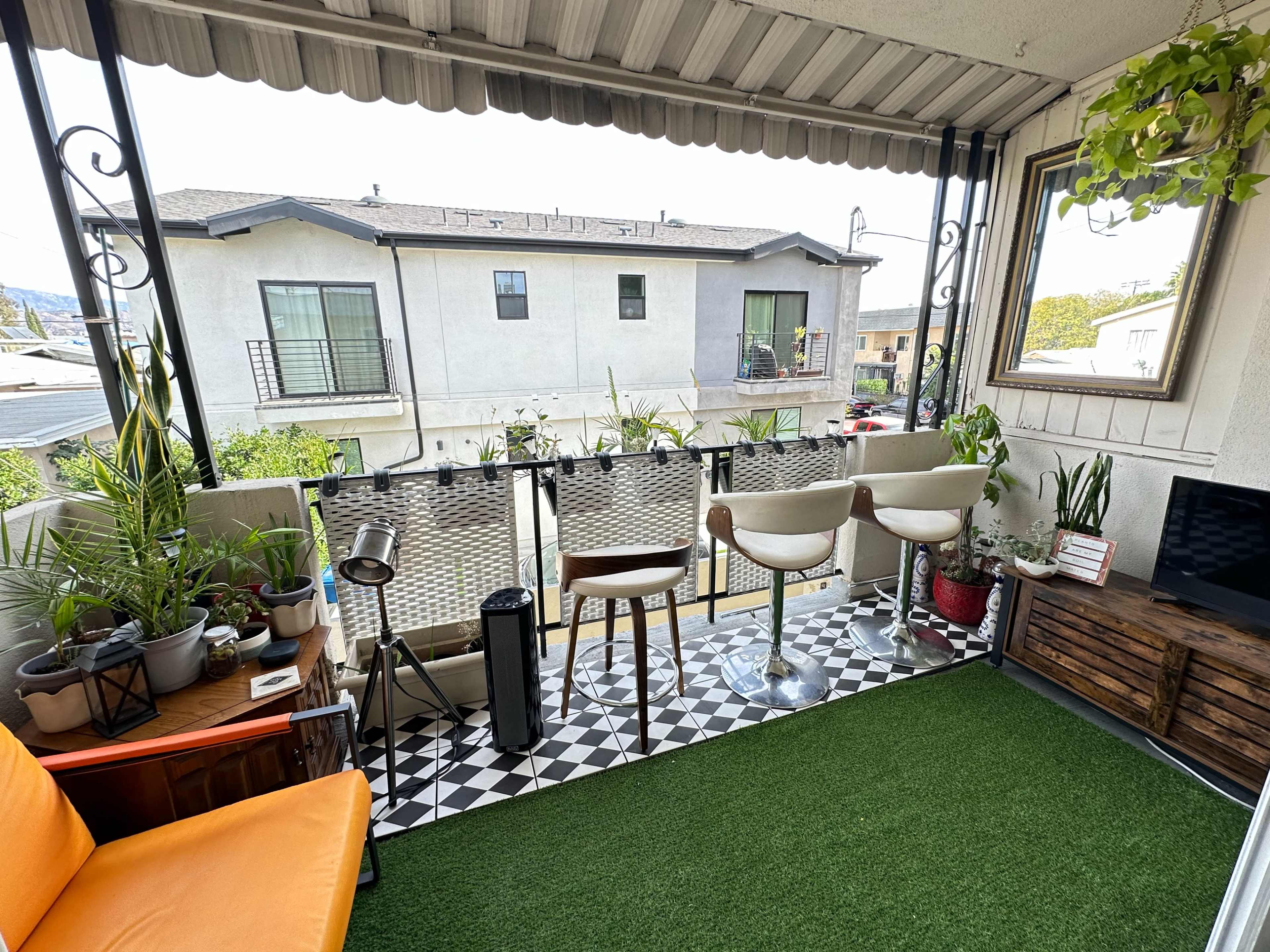 A small balcony with two bar stools, potted plants, and a grassy mat, overlooking neighboring buildings.