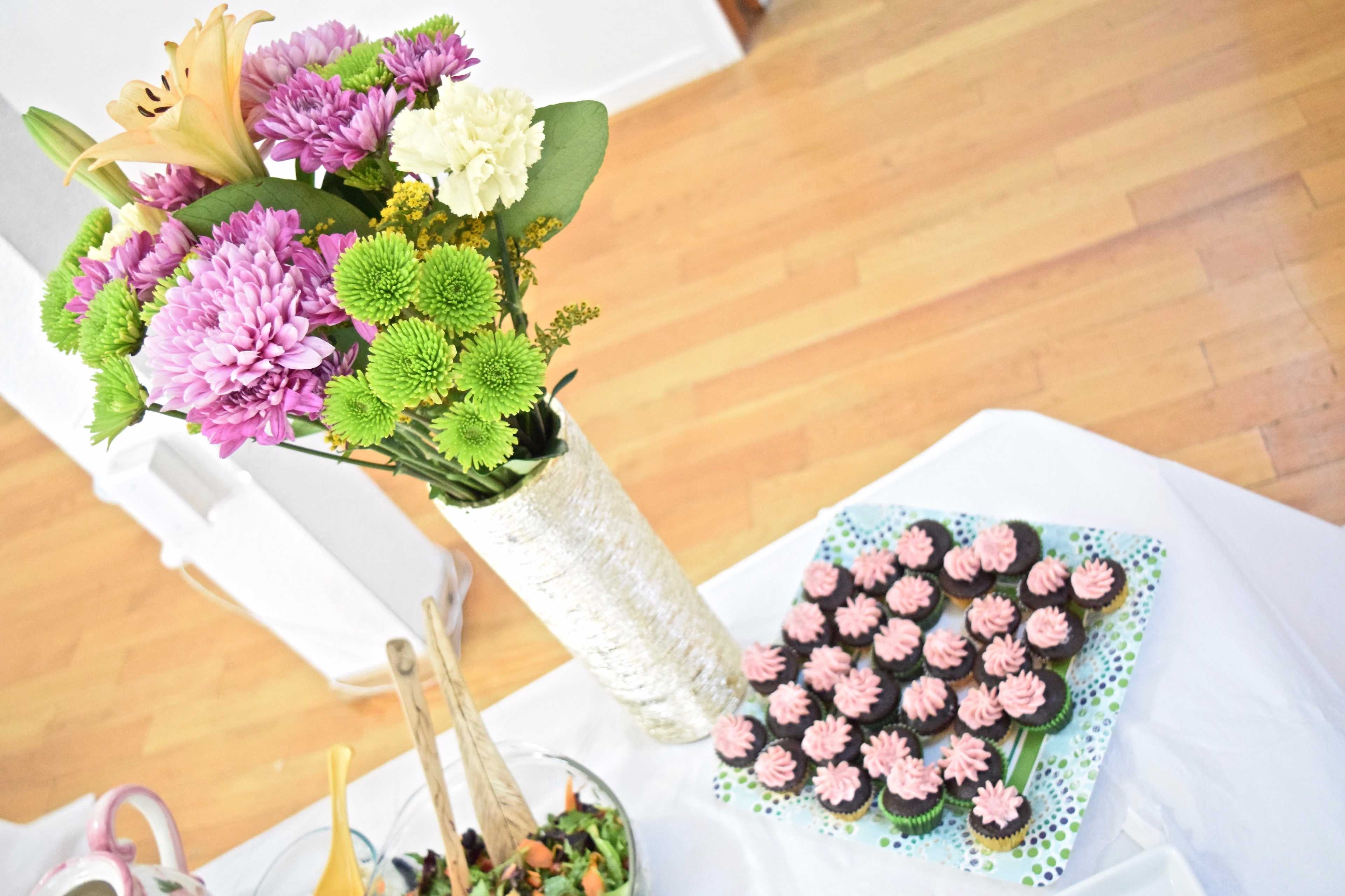 A vase of flowers next to a platter of cupcakes on a table with a wooden floor.