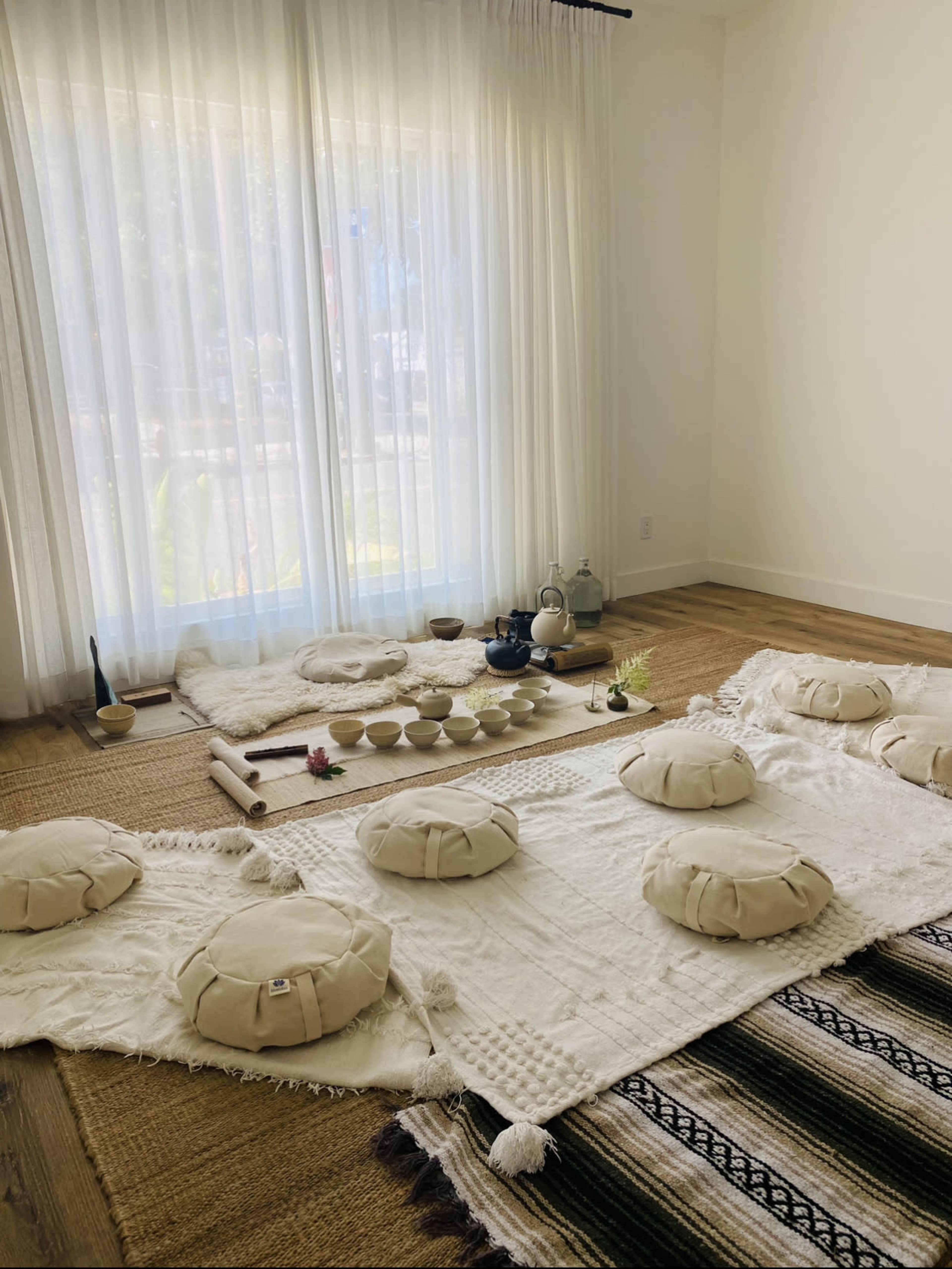A minimalist meditation space with several round cushions on woven rugs, arranged around a low table set with bowls and decorative items, in front of large, light-filtering curtains.