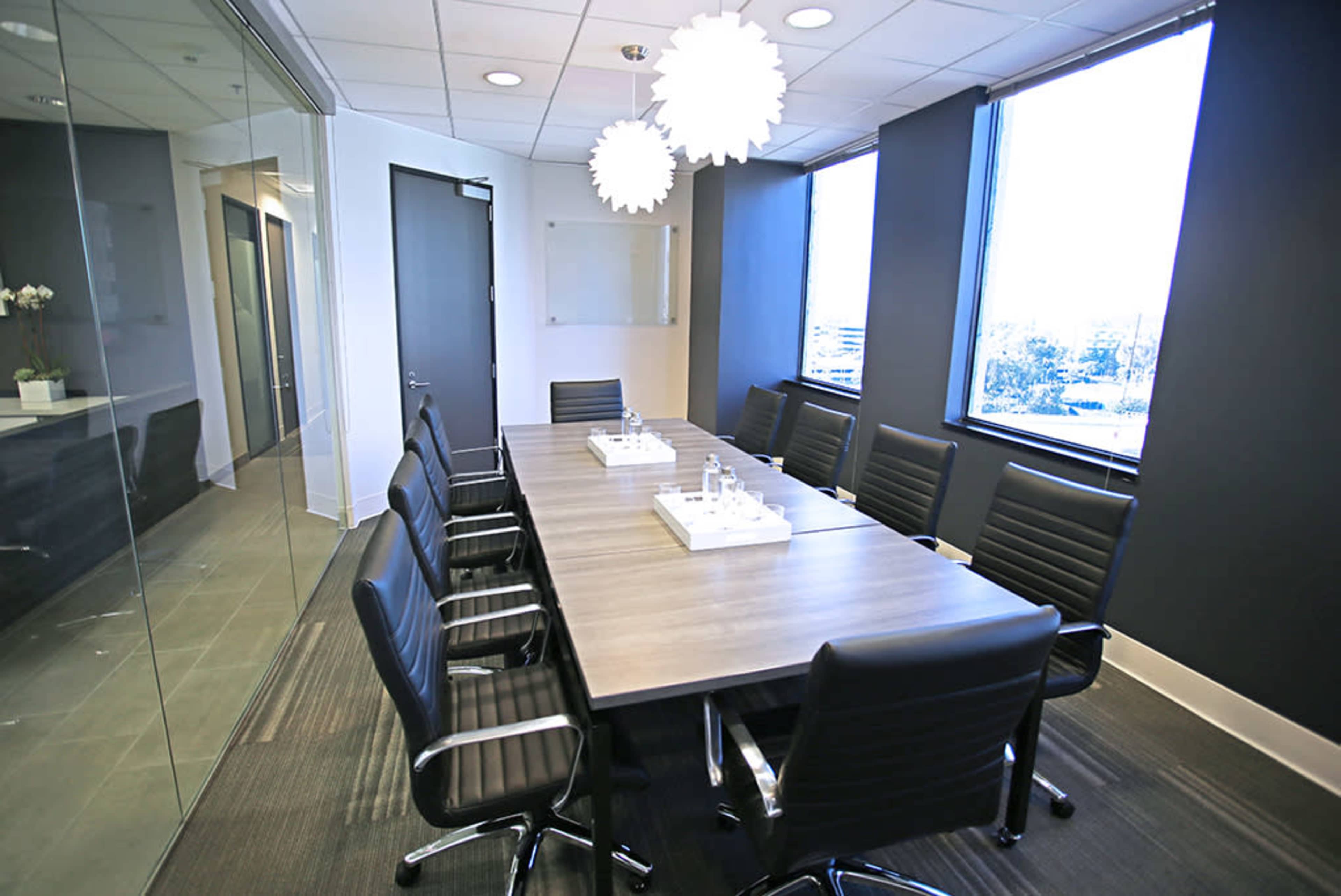 A modern conference room features a large wooden table surrounded by black chairs, with large windows offering a view of the outside.