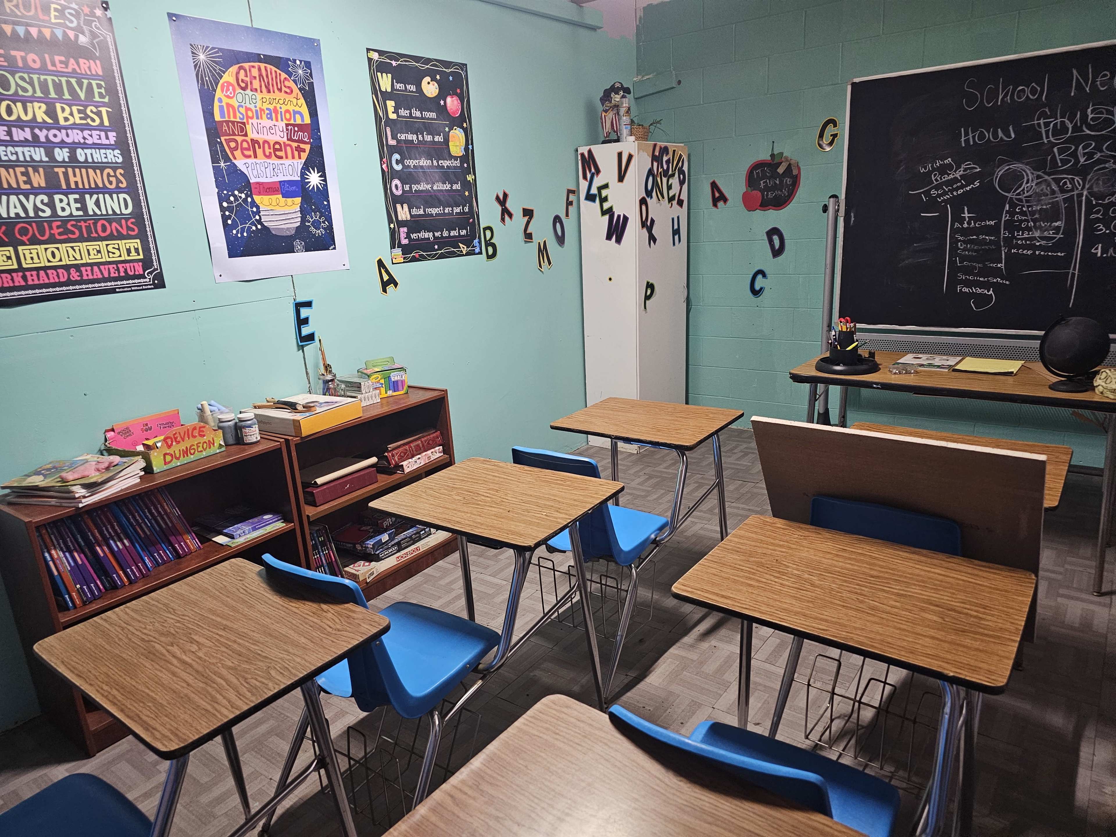 A classroom is arranged with several blue chairs and wooden desks, featuring educational posters on the walls and a chalkboard at the front.