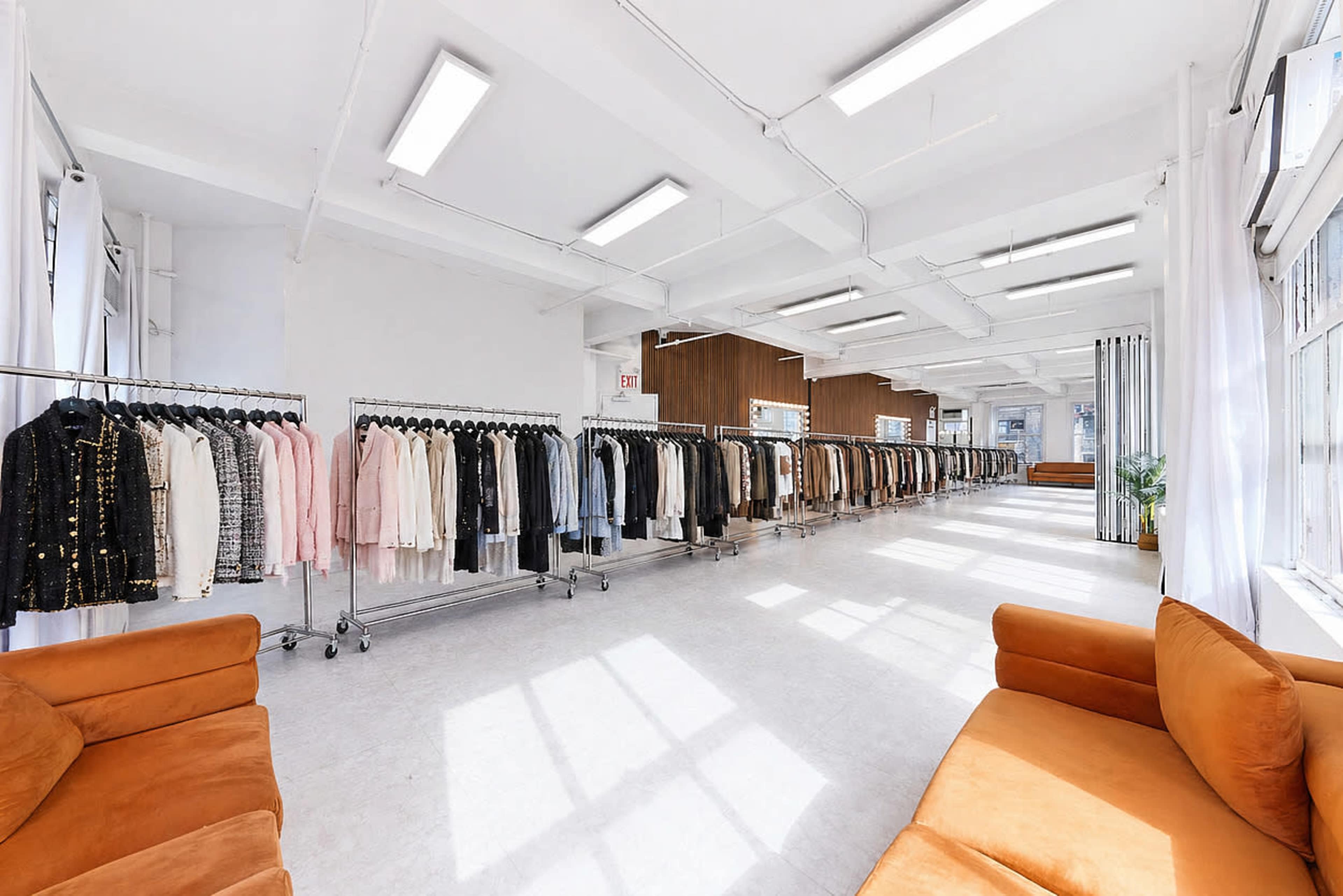 The image shows a spacious clothing store interior with racks of various garments lined along one side and two orange sofas arranged in the center.