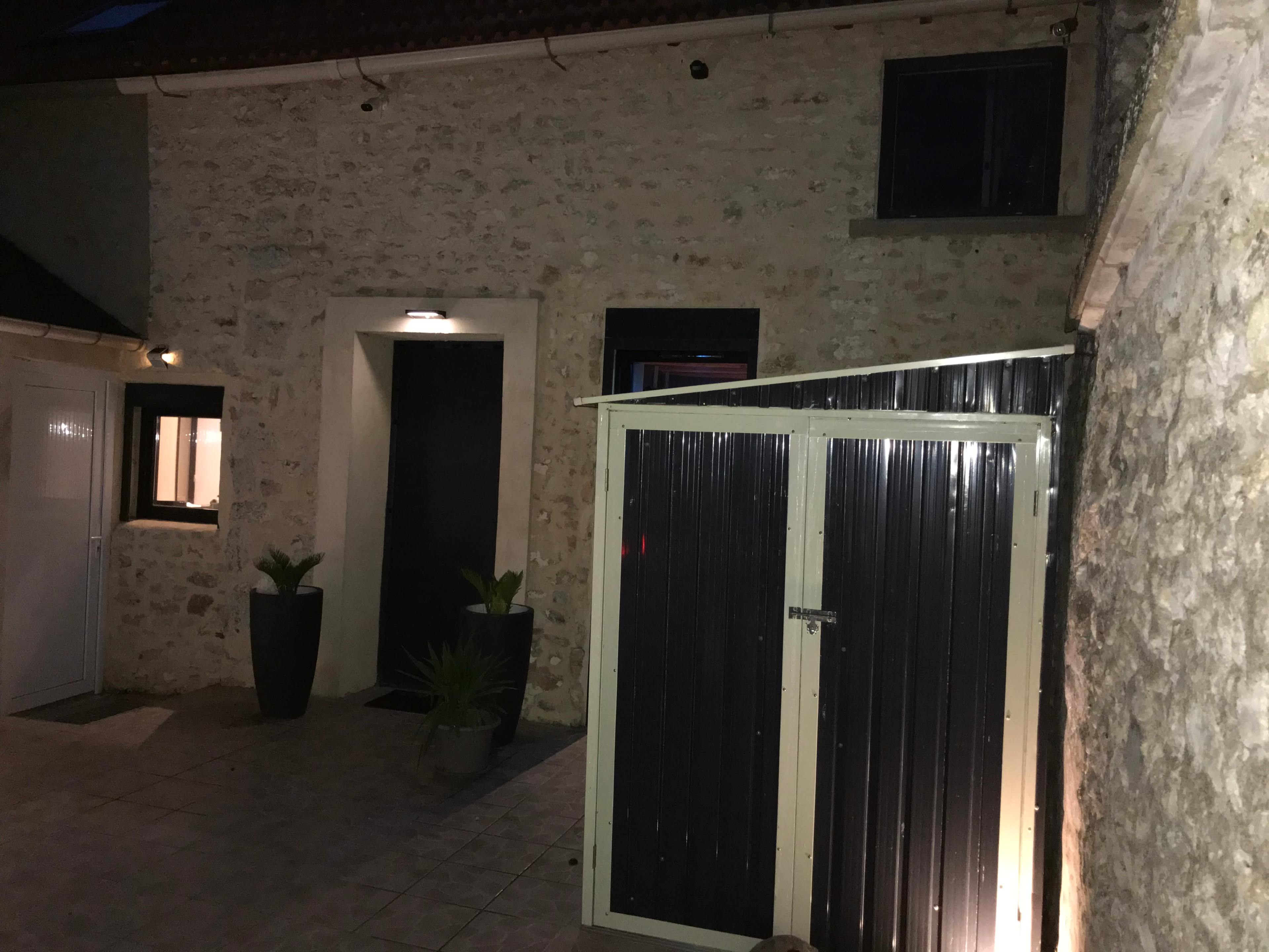 A stone building with a dark doorway, two windows, and a metal storage shed beside it, illuminated at night.