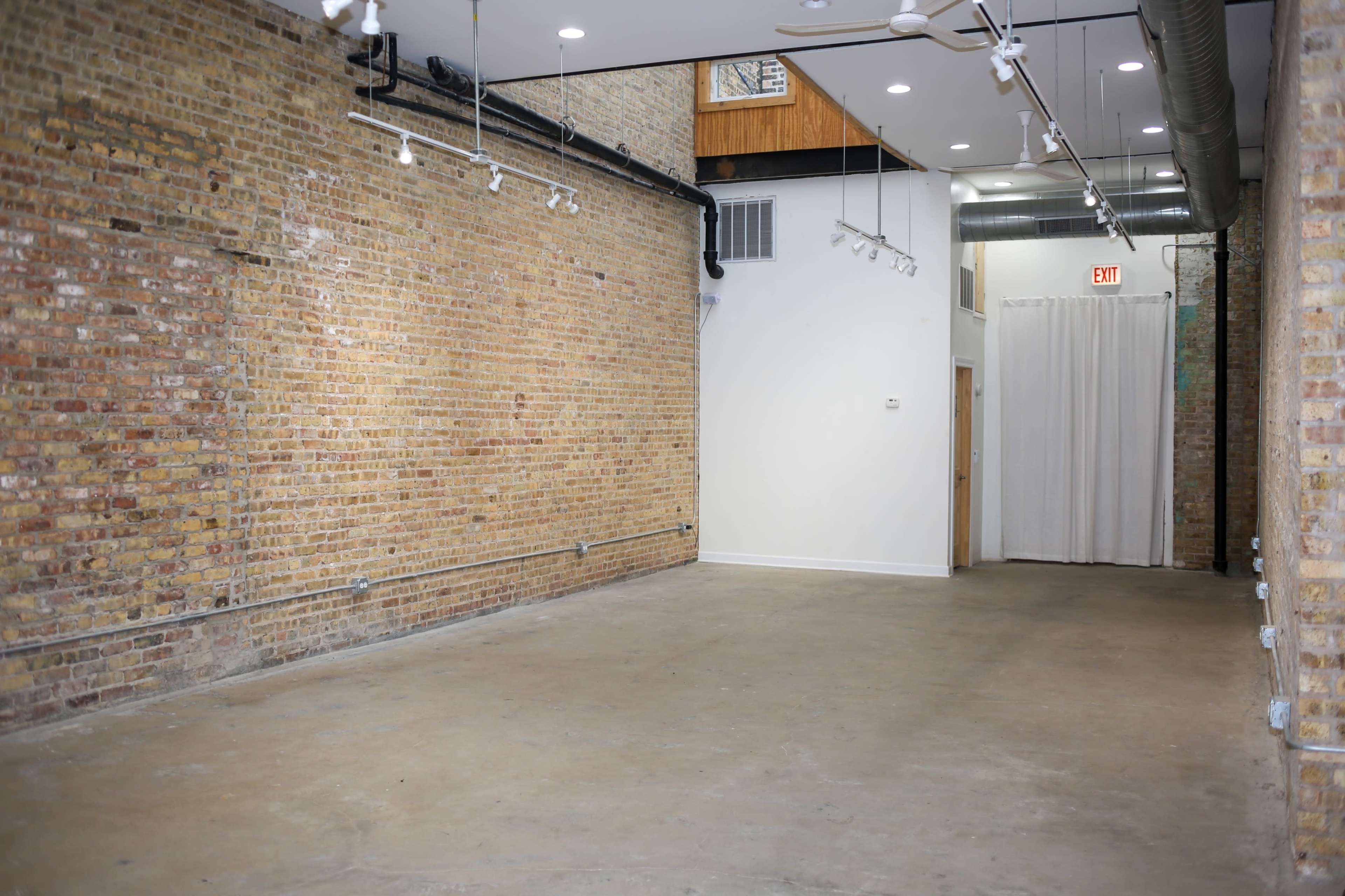 An empty interior space with exposed brick walls and a concrete floor, well-lit by overhead track lighting.
