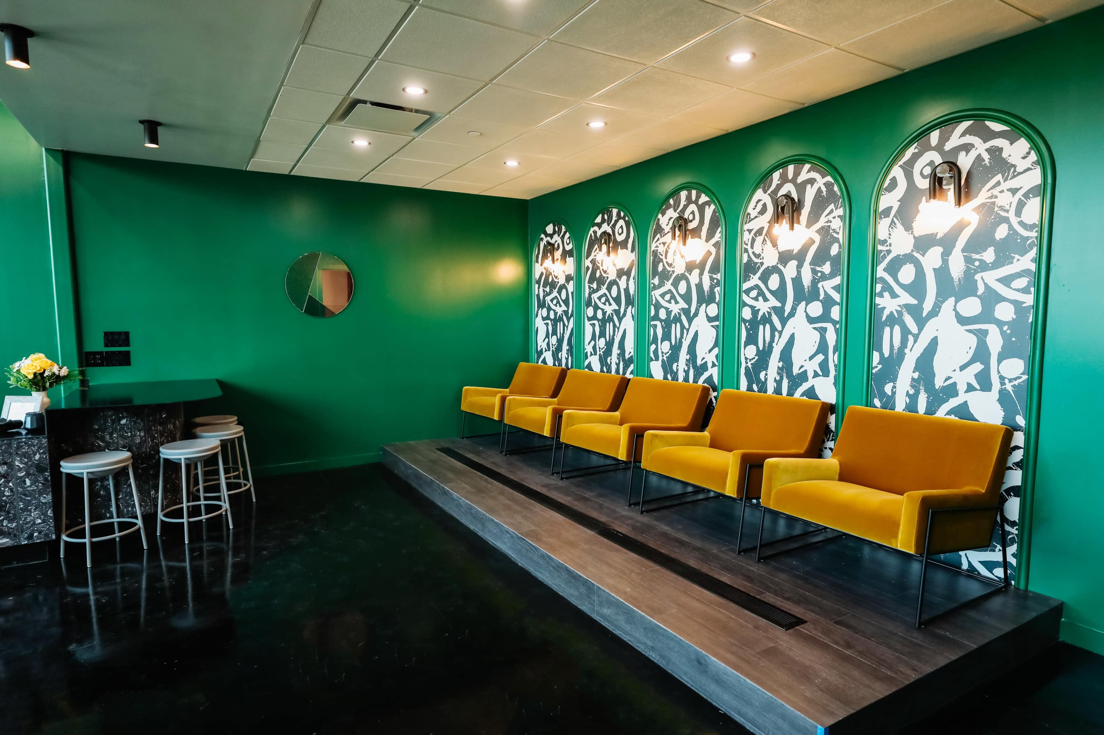 The image shows a waiting area with bright green walls, a patterned mural, four yellow chairs, and a bar with stools against one wall.