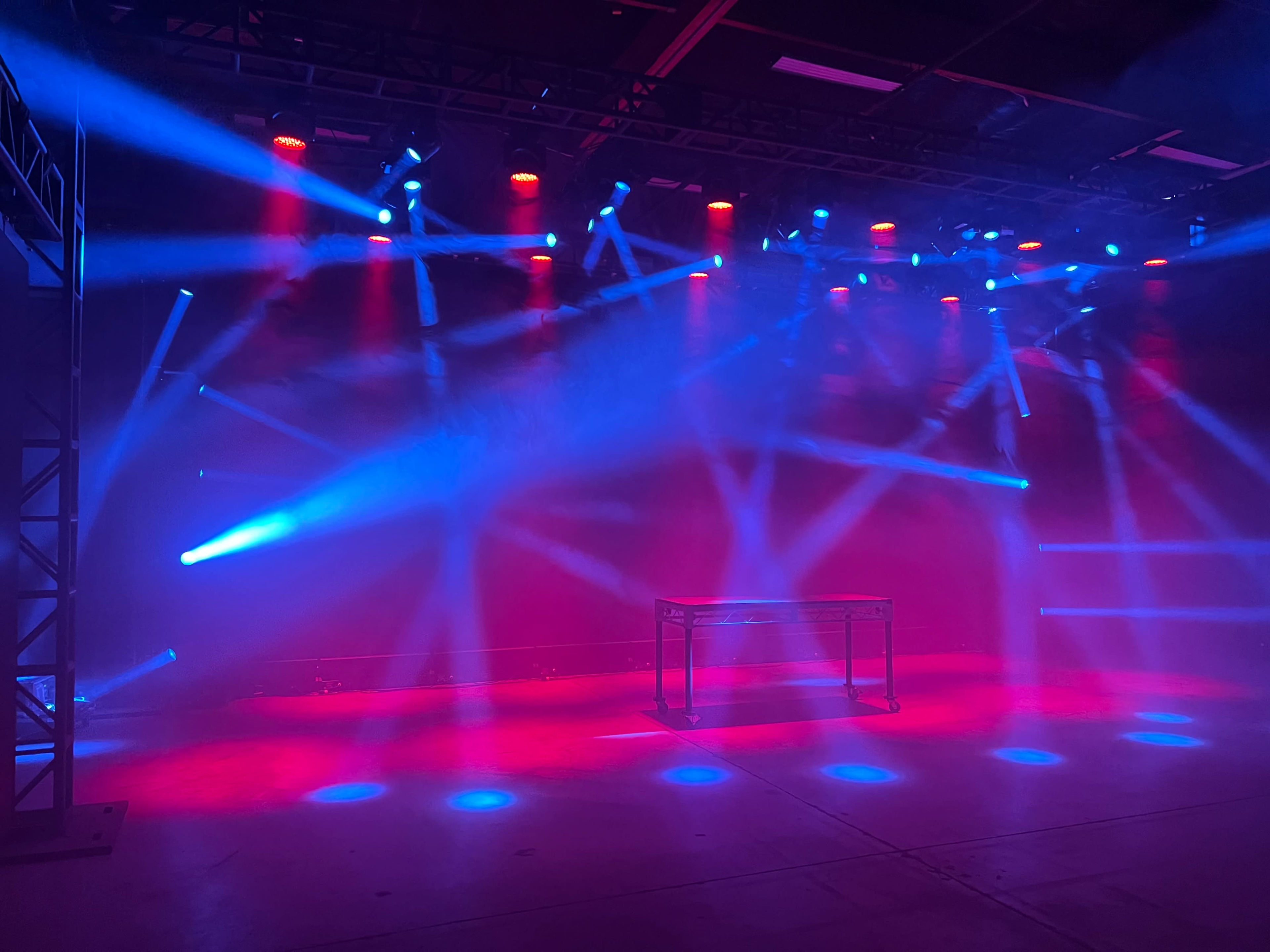 A stage setup with vibrant blue and red lighting illuminating a metallic table under various beams of light in a misty environment.