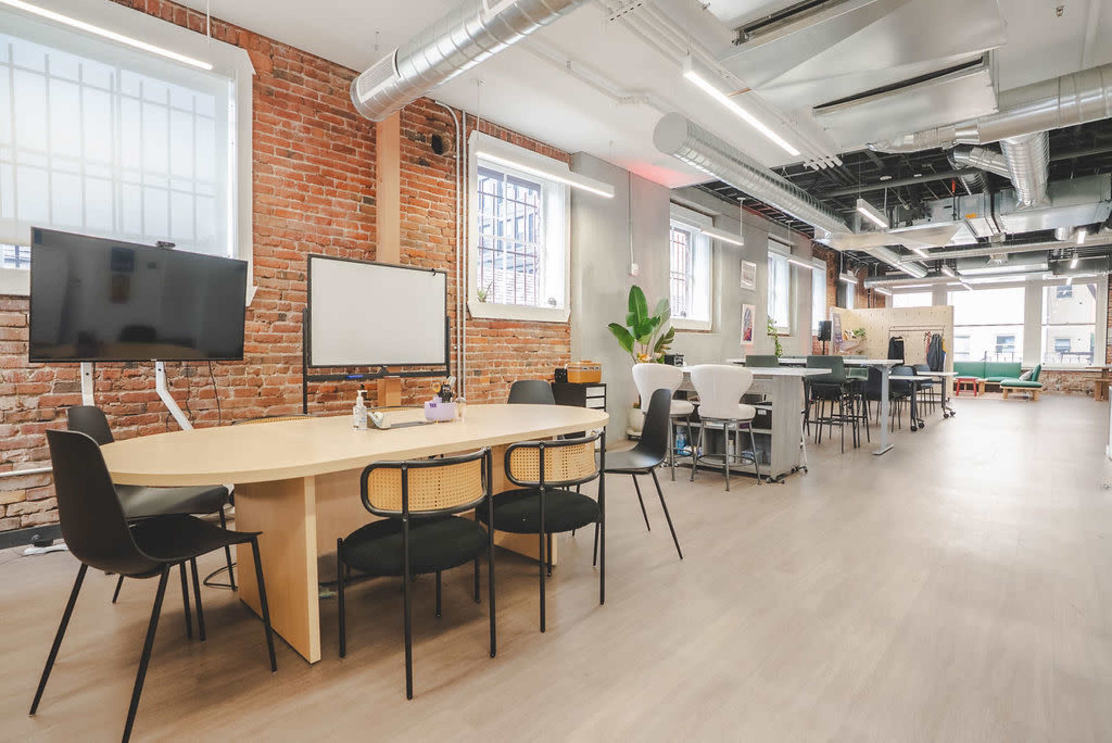 The image shows an open office space featuring a large wooden table, black chairs, brick walls, and various workstations with modern furnishings.