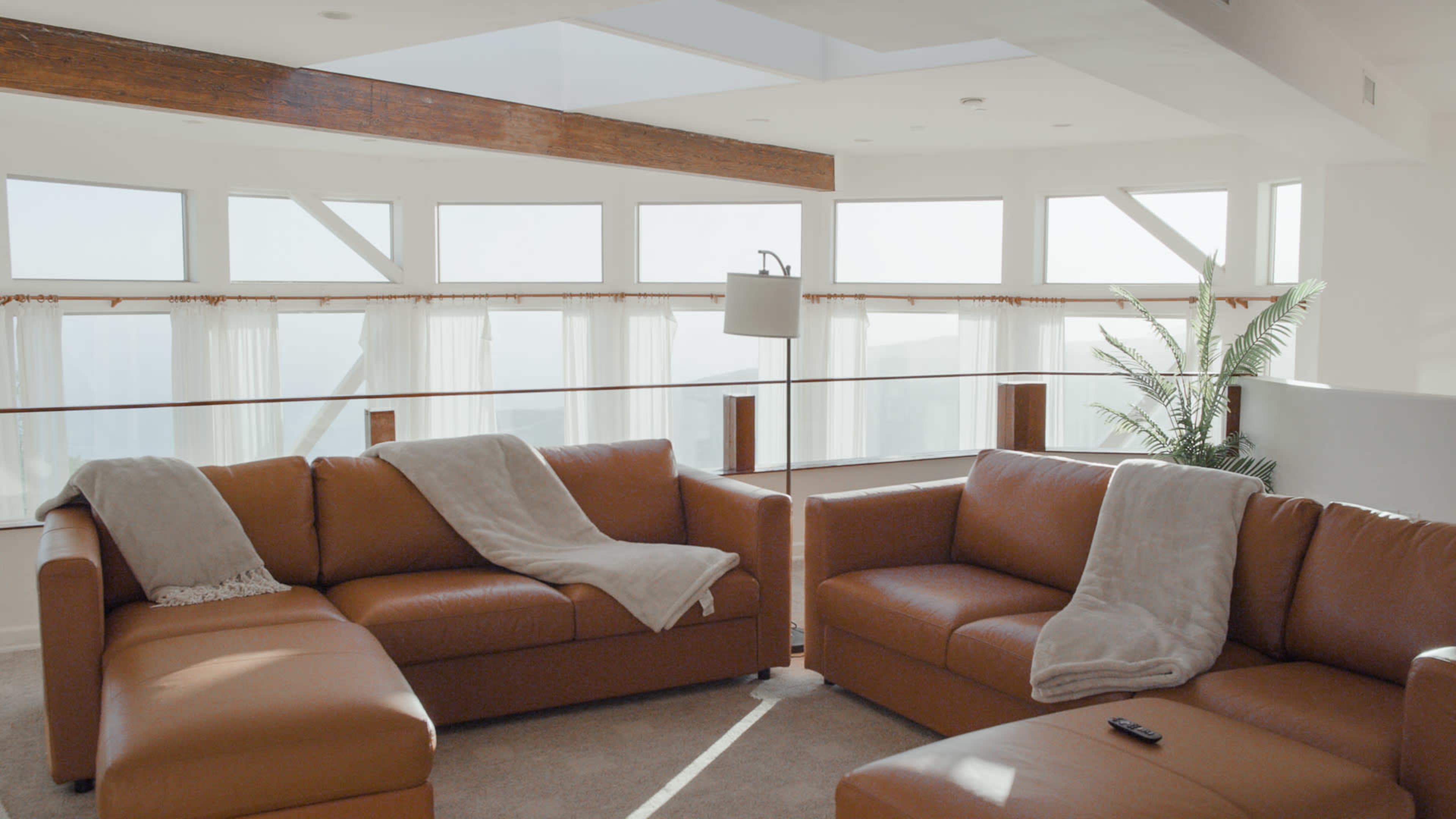 A cozy living room with large windows, featuring a sectional brown leather sofa and a floor lamp.
