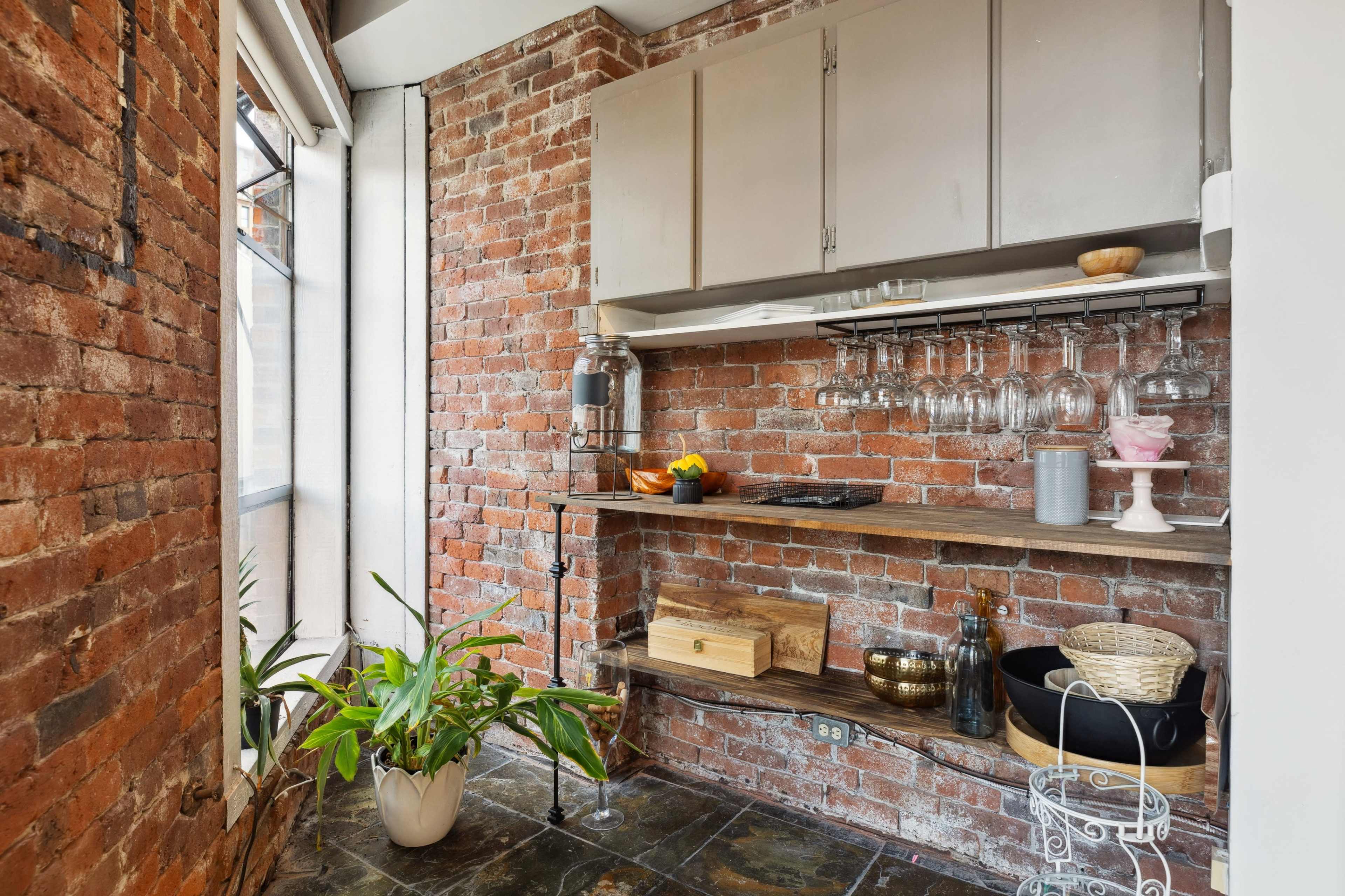 A modern kitchen area featuring exposed brick walls, wooden shelves with various kitchen items, and a potted plant.