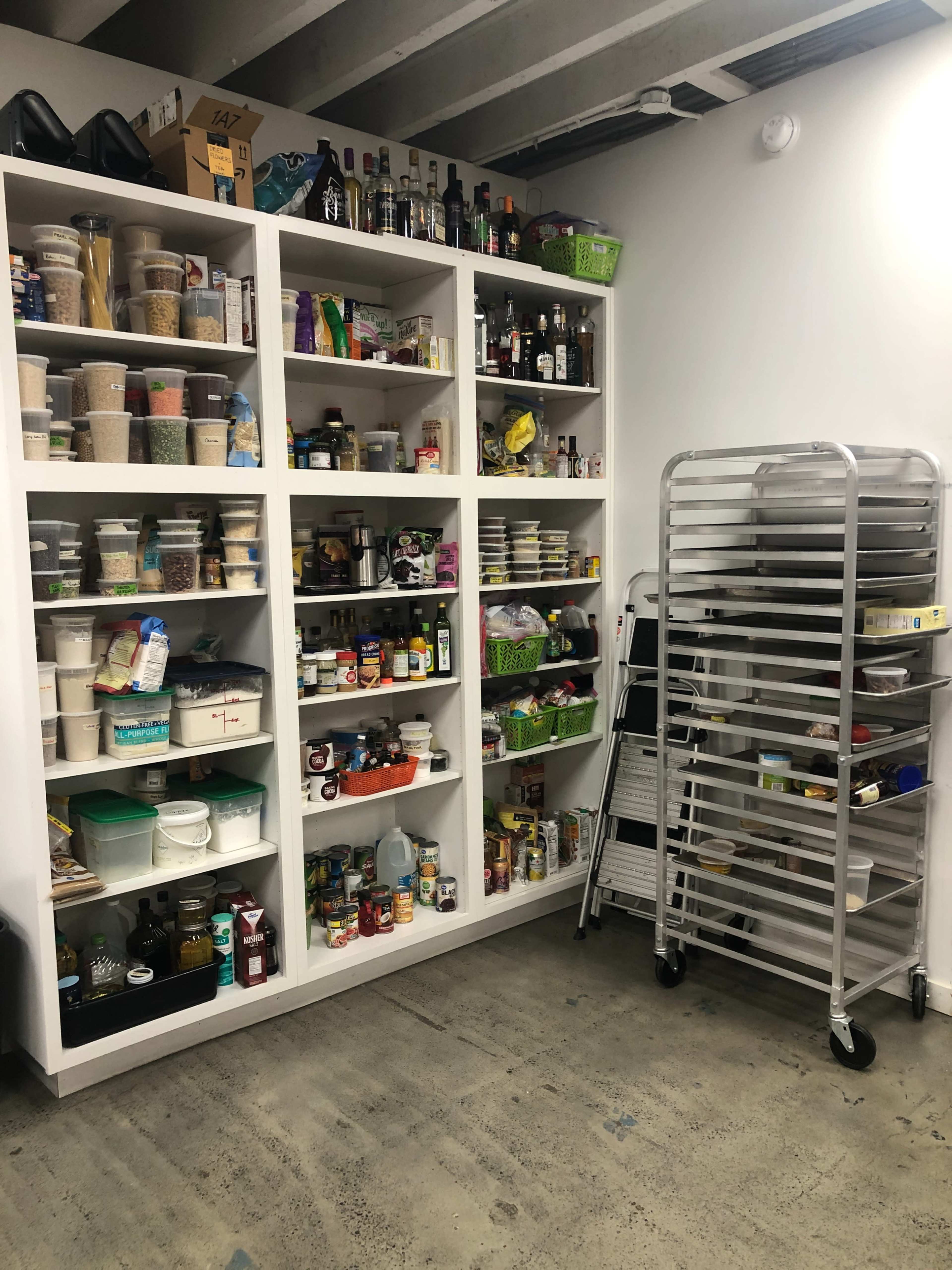A storage area with shelves filled with various jars, cans, and bottles, alongside a mobile rack in a corner.