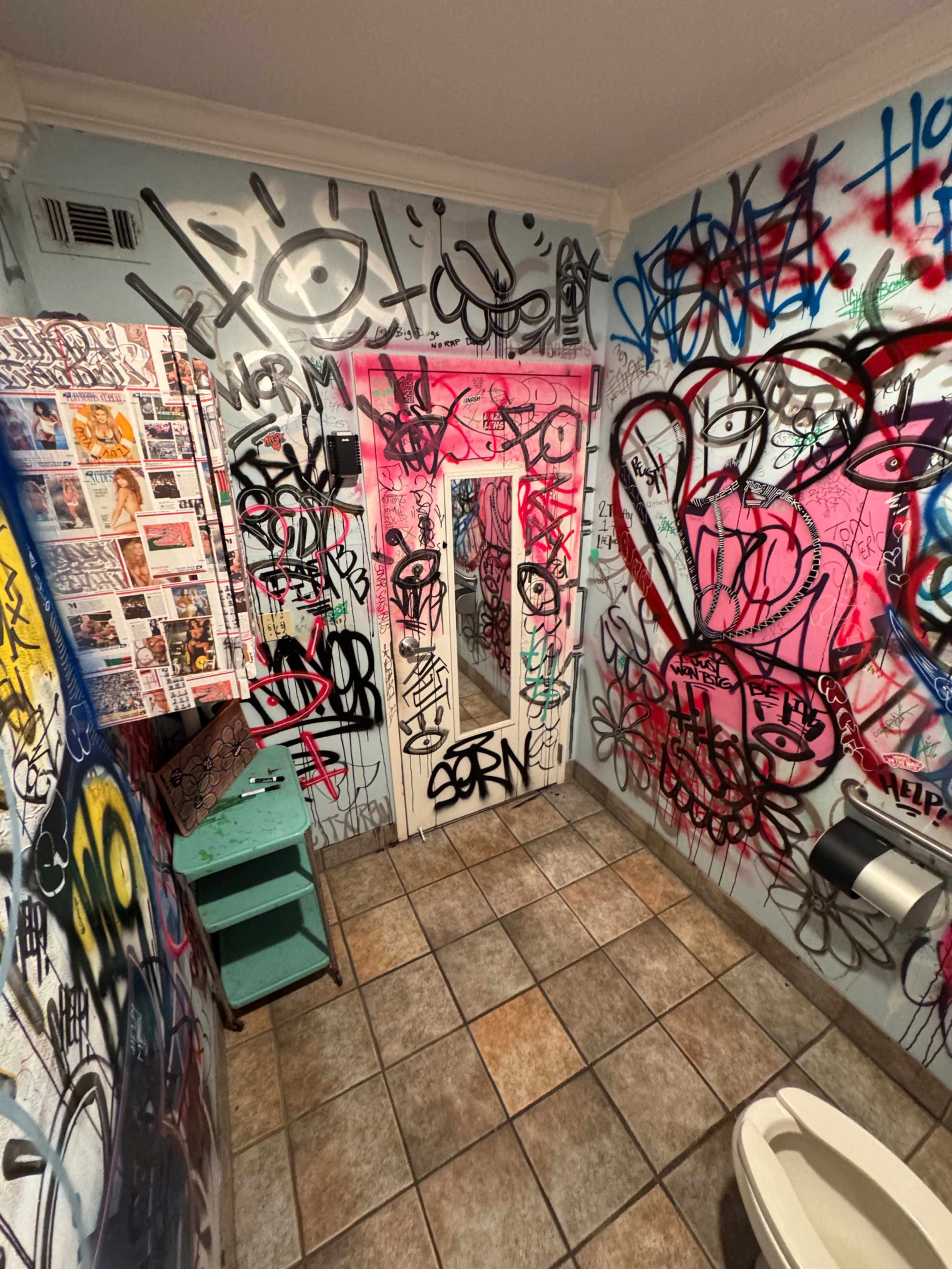 The bathroom features walls covered in vibrant graffiti, with a tiled floor and a door leading to another area.