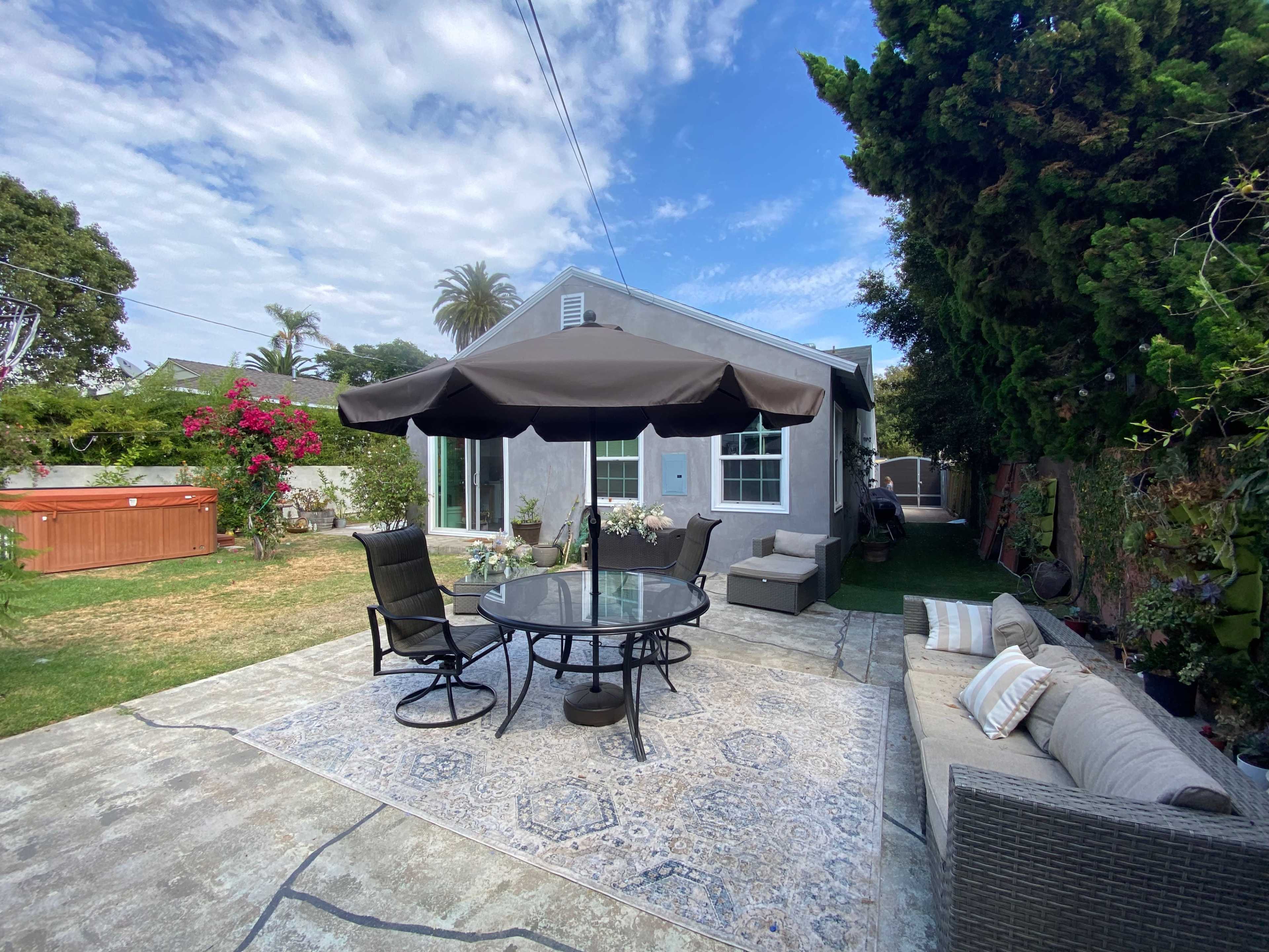 A small backyard features a round table with chairs under a large umbrella, a hot tub, and a gray house surrounded by greenery.