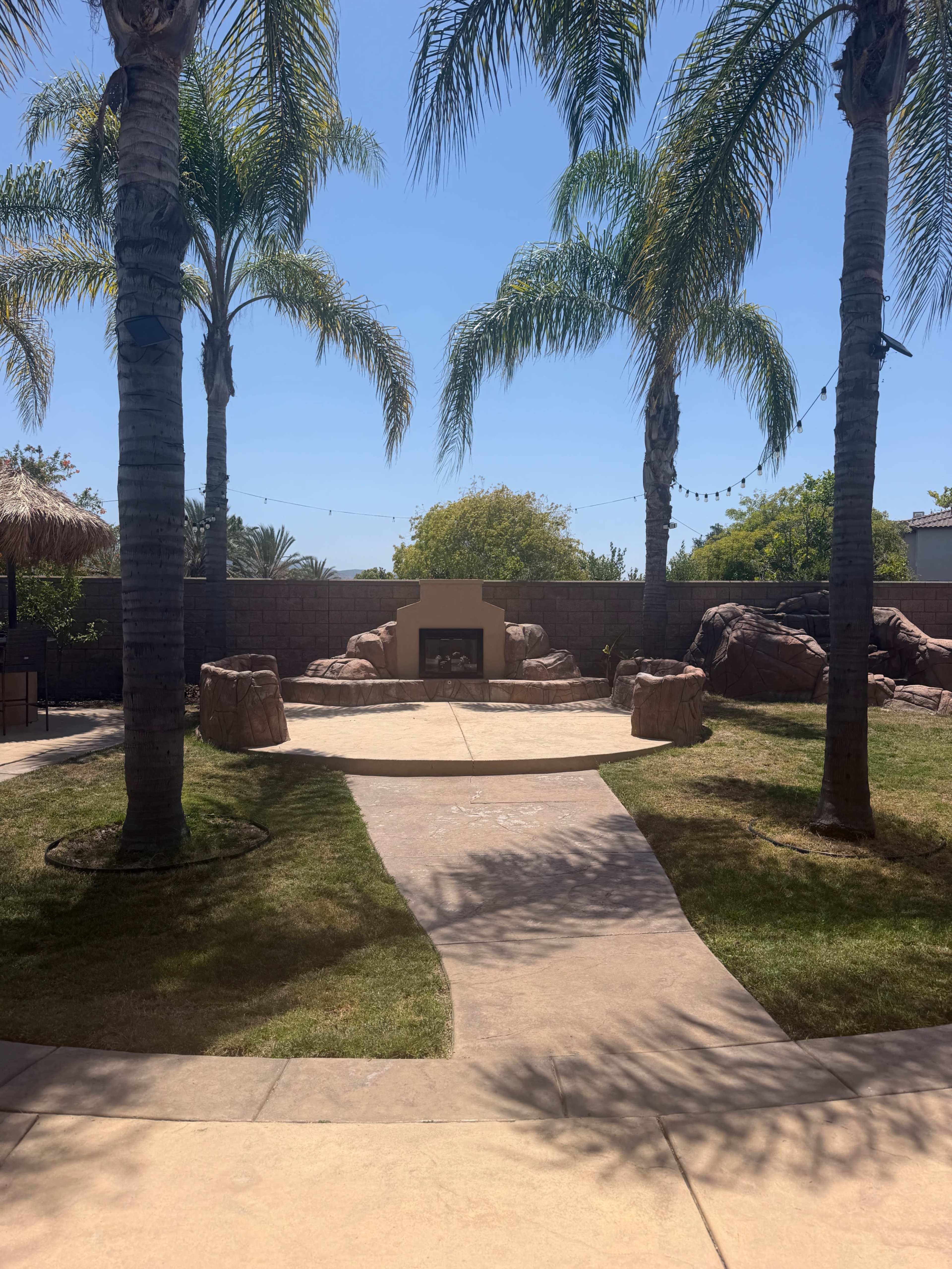 The image shows a landscaped outdoor space with a stone fireplace surrounded by palm trees and a concrete path leading to it.