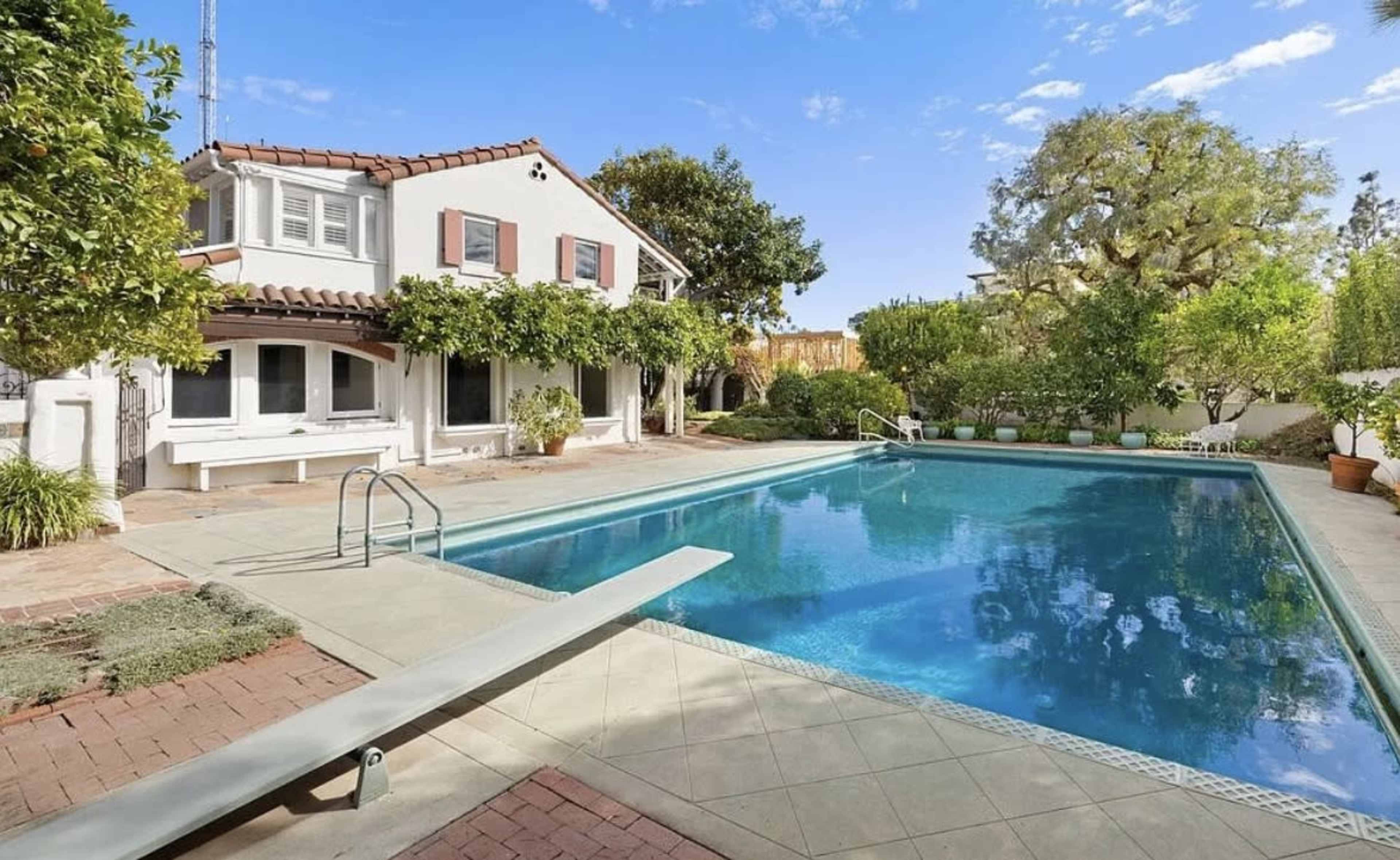 The image shows a residential backyard with a swimming pool, surrounded by greenery and a two-story house with a patio area.