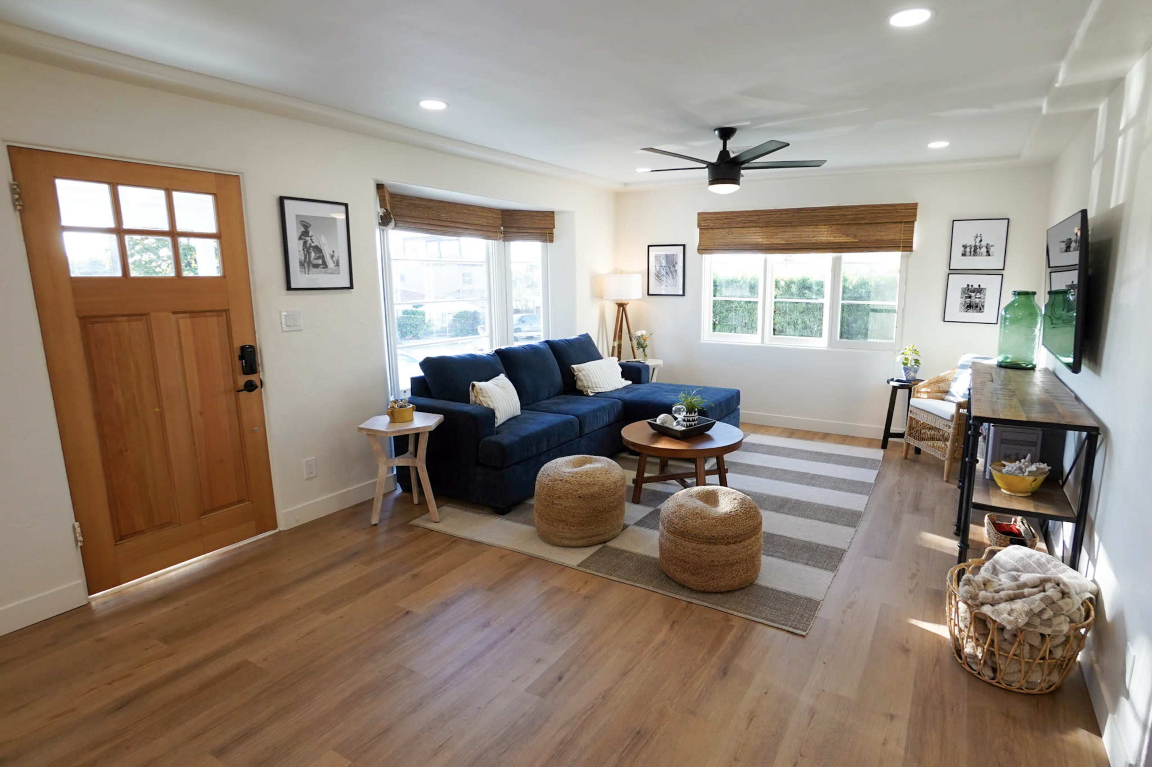 The living room features a navy blue sofa, a wooden coffee table, and two round ottomans, with large windows allowing natural light and a front door made of wood.