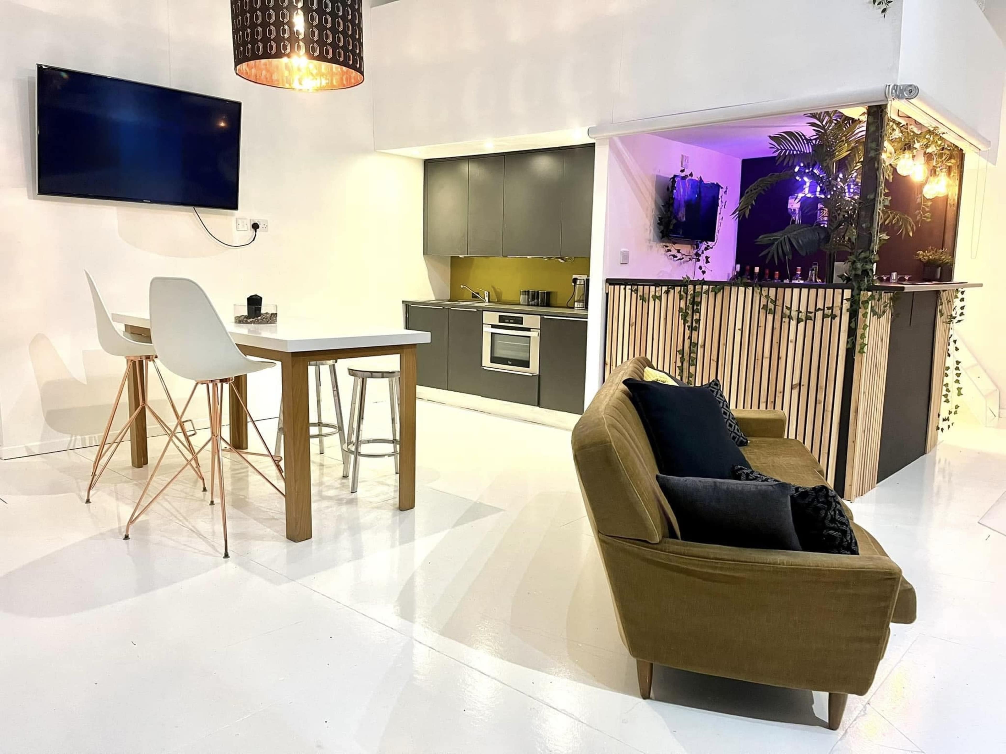 The image shows a modern, open-concept living space featuring a kitchen area, a dining table with chairs, a couch, and a television mounted on the wall.