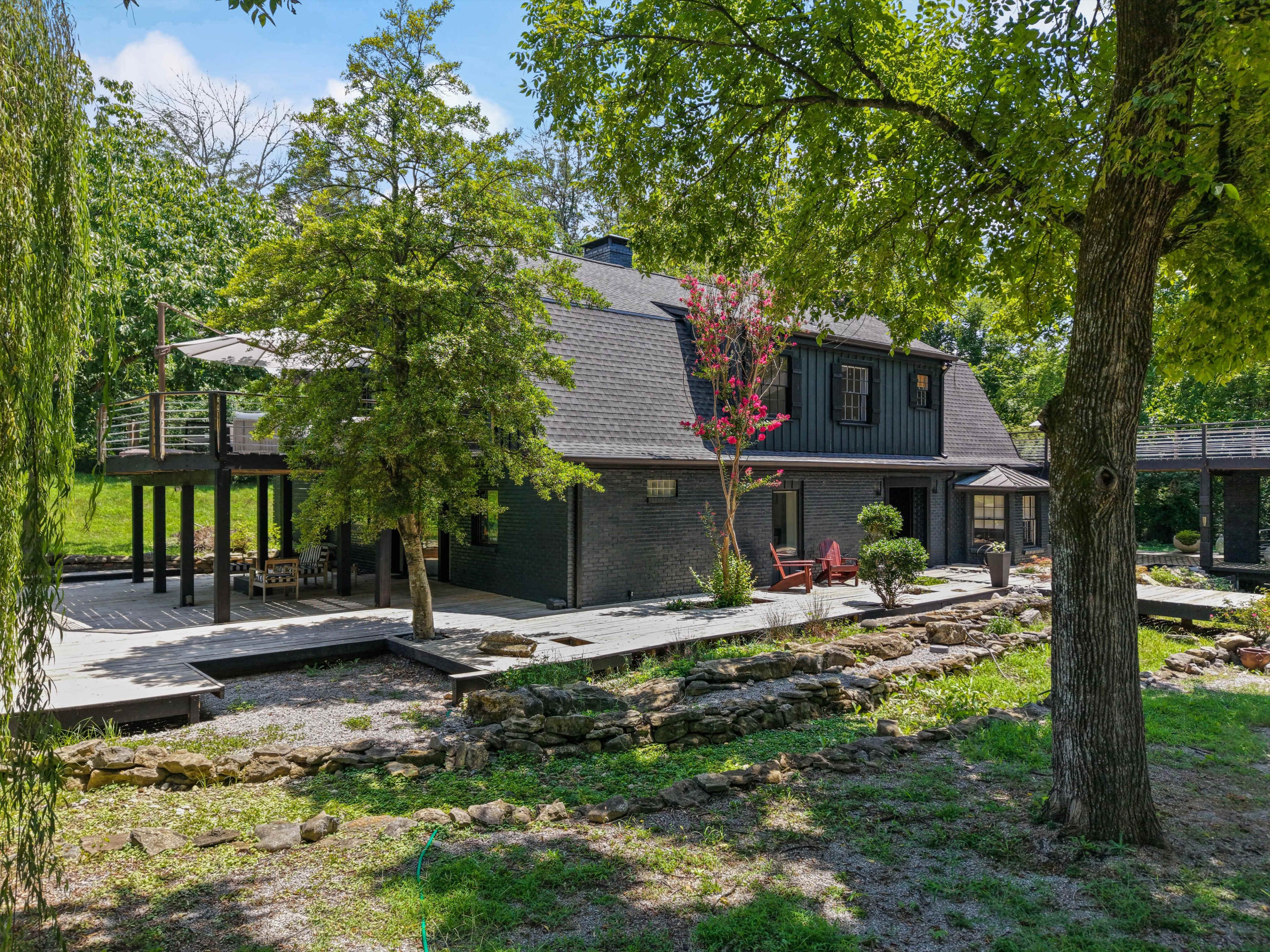 The image shows a two-story black wooden house surrounded by greenery and a deck area with seating.