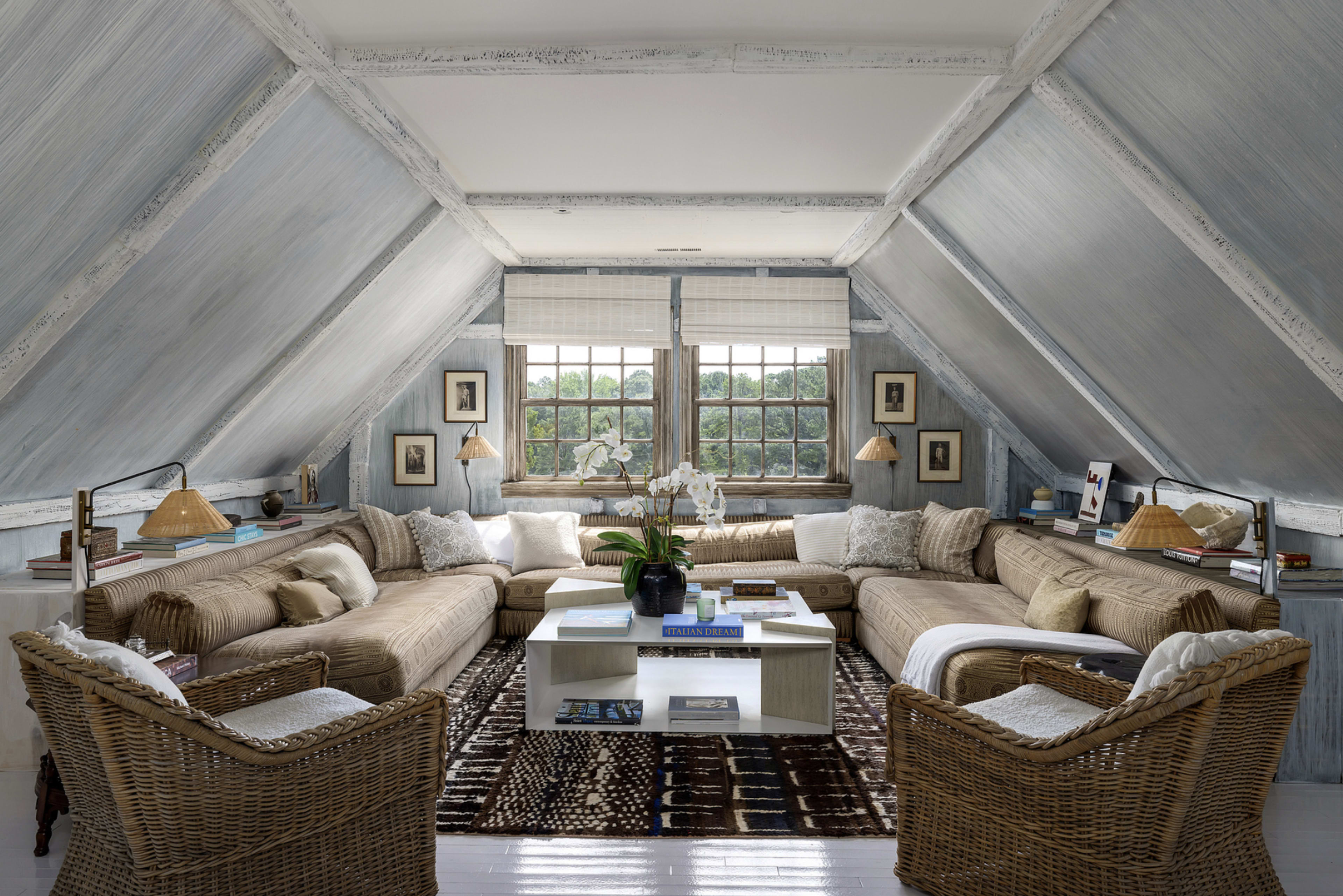 The image shows a cozy attic living area with two large sectional sofas, a coffee table, and decorative artwork on the walls, illuminated by natural light from large windows.