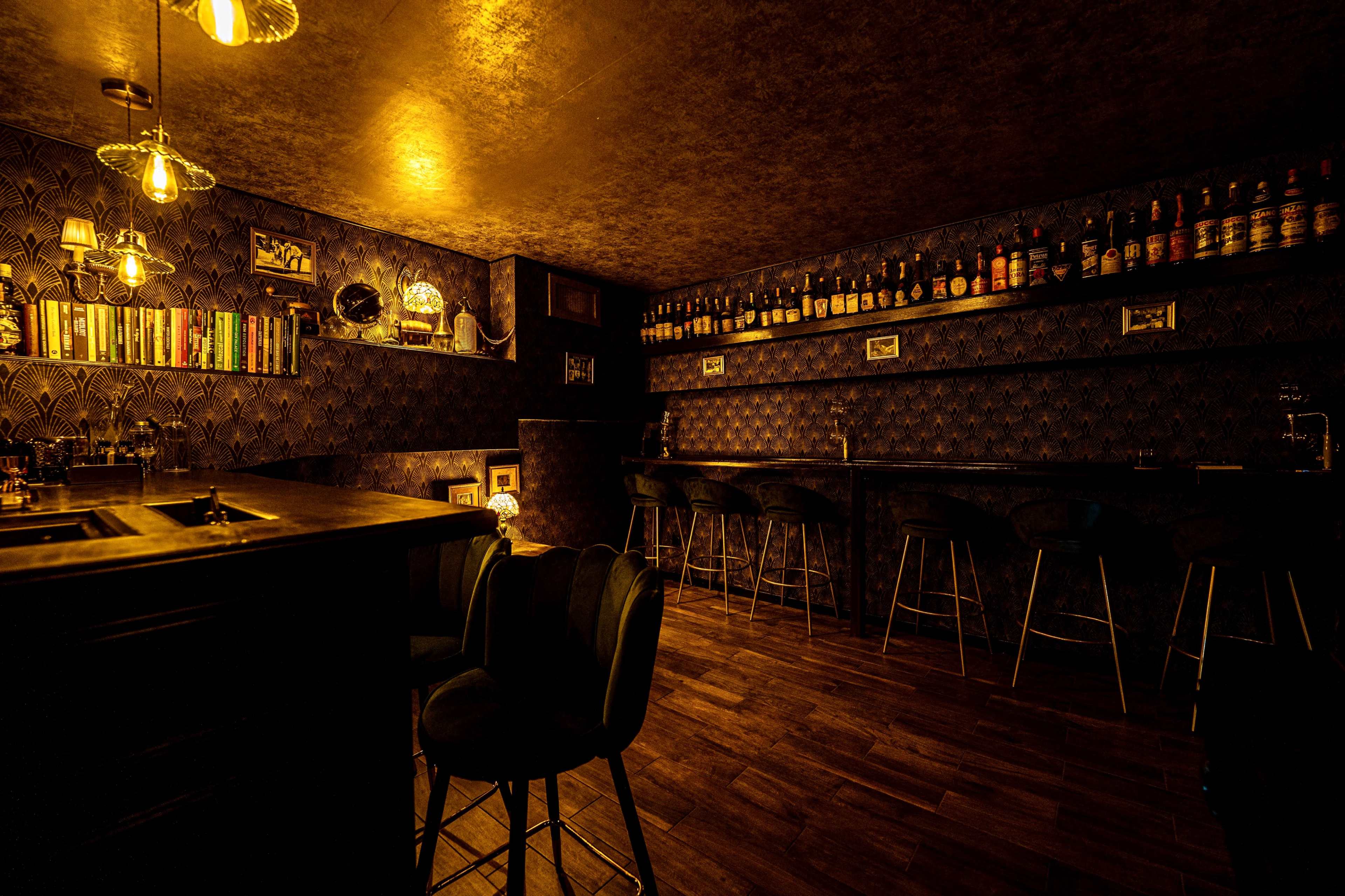 The image depicts a dimly lit bar with wooden flooring, a dark decor featuring shelves of bottles, and bar stools arranged around a counter.