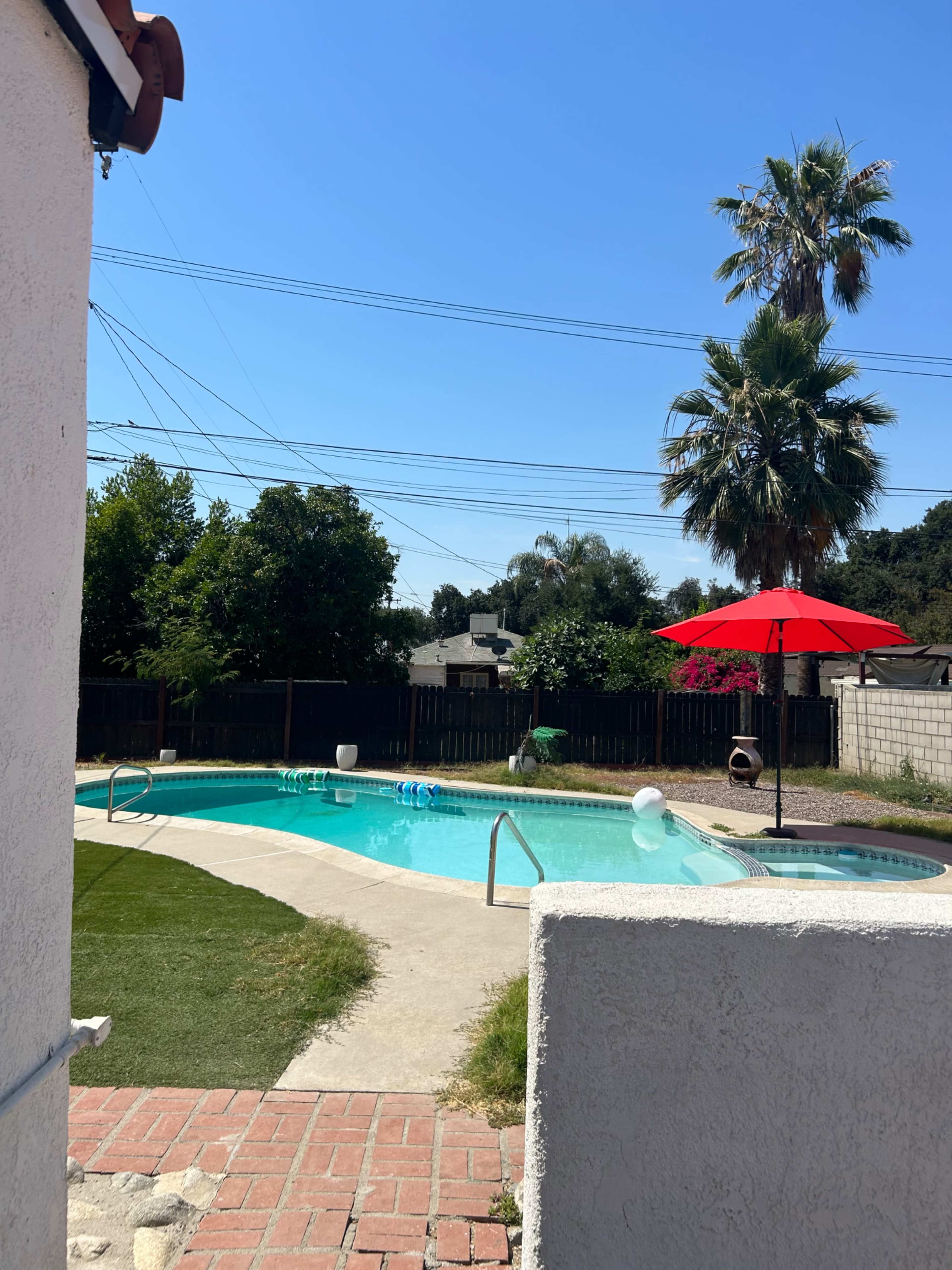 A patio area overlooks a swimming pool surrounded by grass and palm trees, with a red umbrella providing shade.