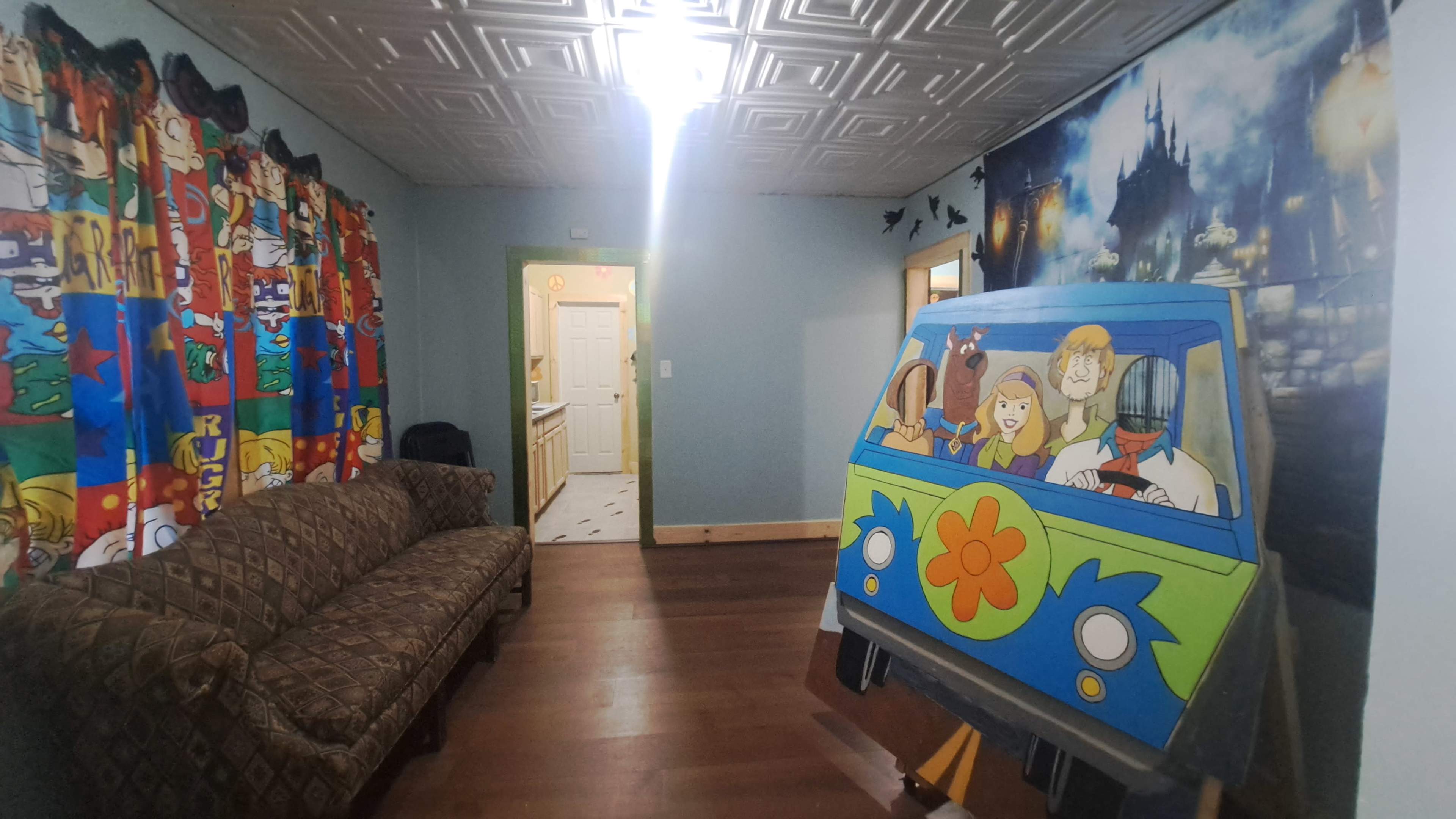 The scene features a colorful room with a cartoon-themed curtain, a patterned sofa, and a mural of a cartoon vehicle accompanied by characters on one wall, with a doorway leading to a kitchen visible in the background.
