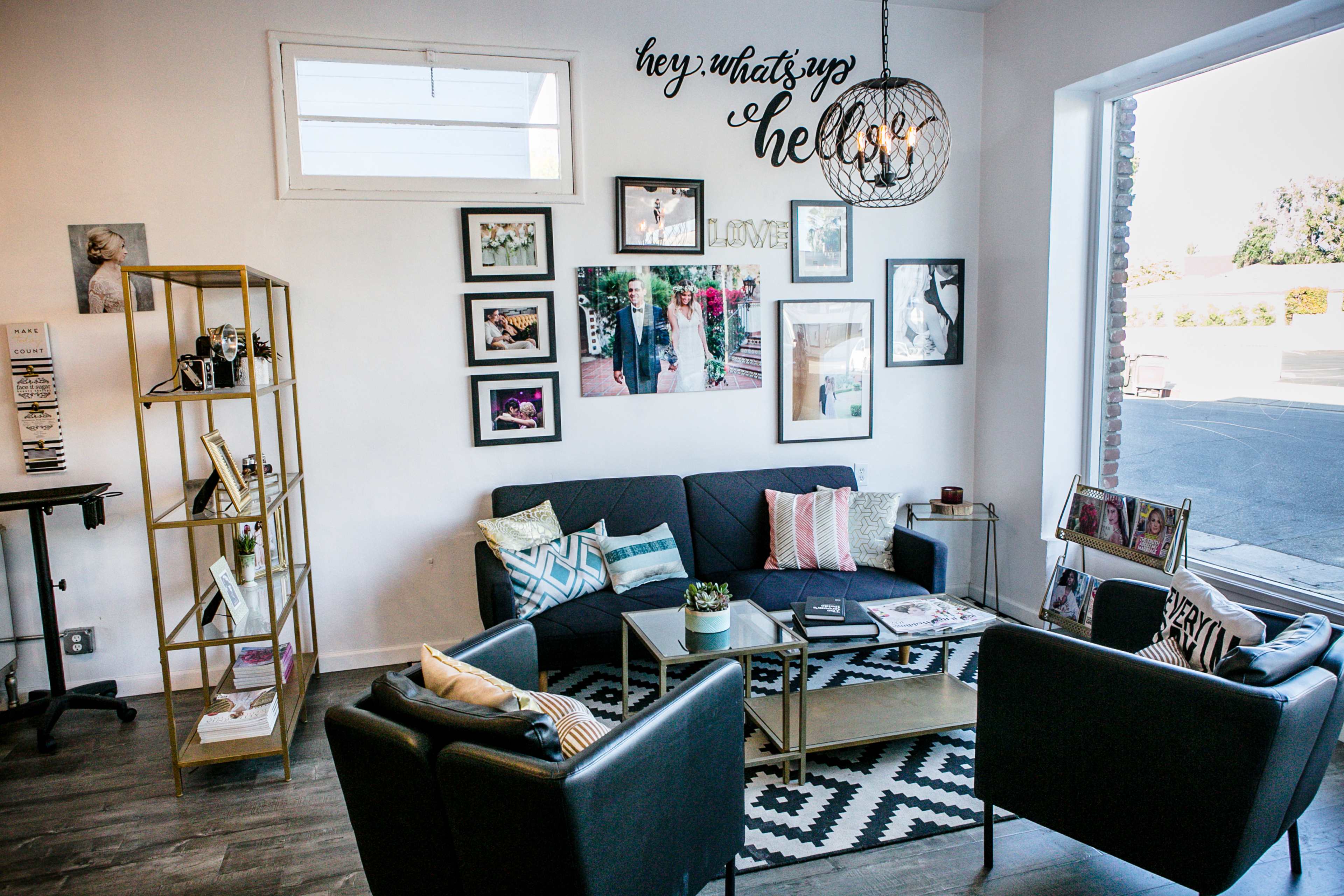 The image shows a modern living room with a black sofa, decorative pillows, a wooden shelving unit, framed photos on the walls, and a light fixture hanging above.