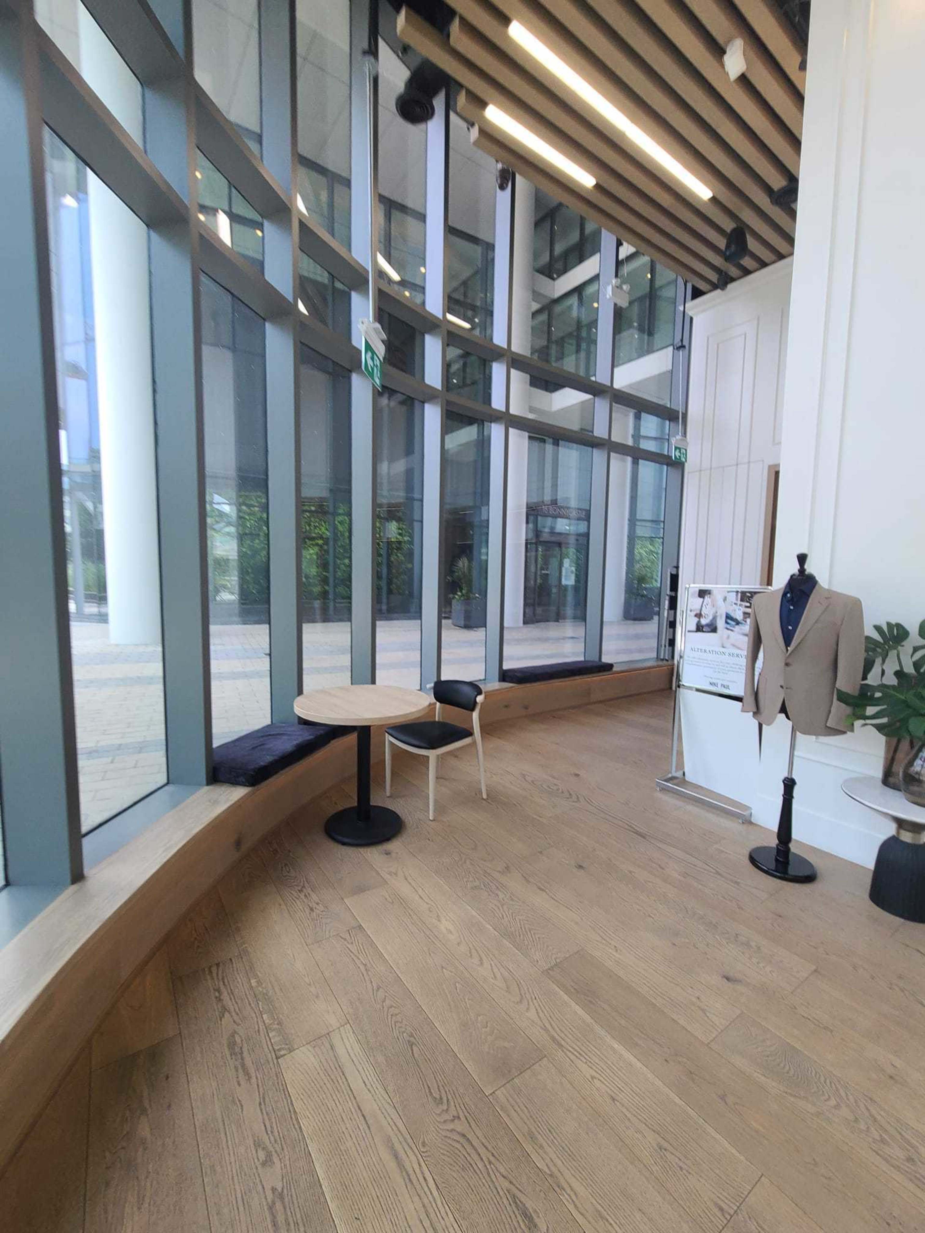 The image shows a modern interior space featuring large glass windows, polished wooden flooring, a round table with a chair, and a display of a suit on a mannequin.