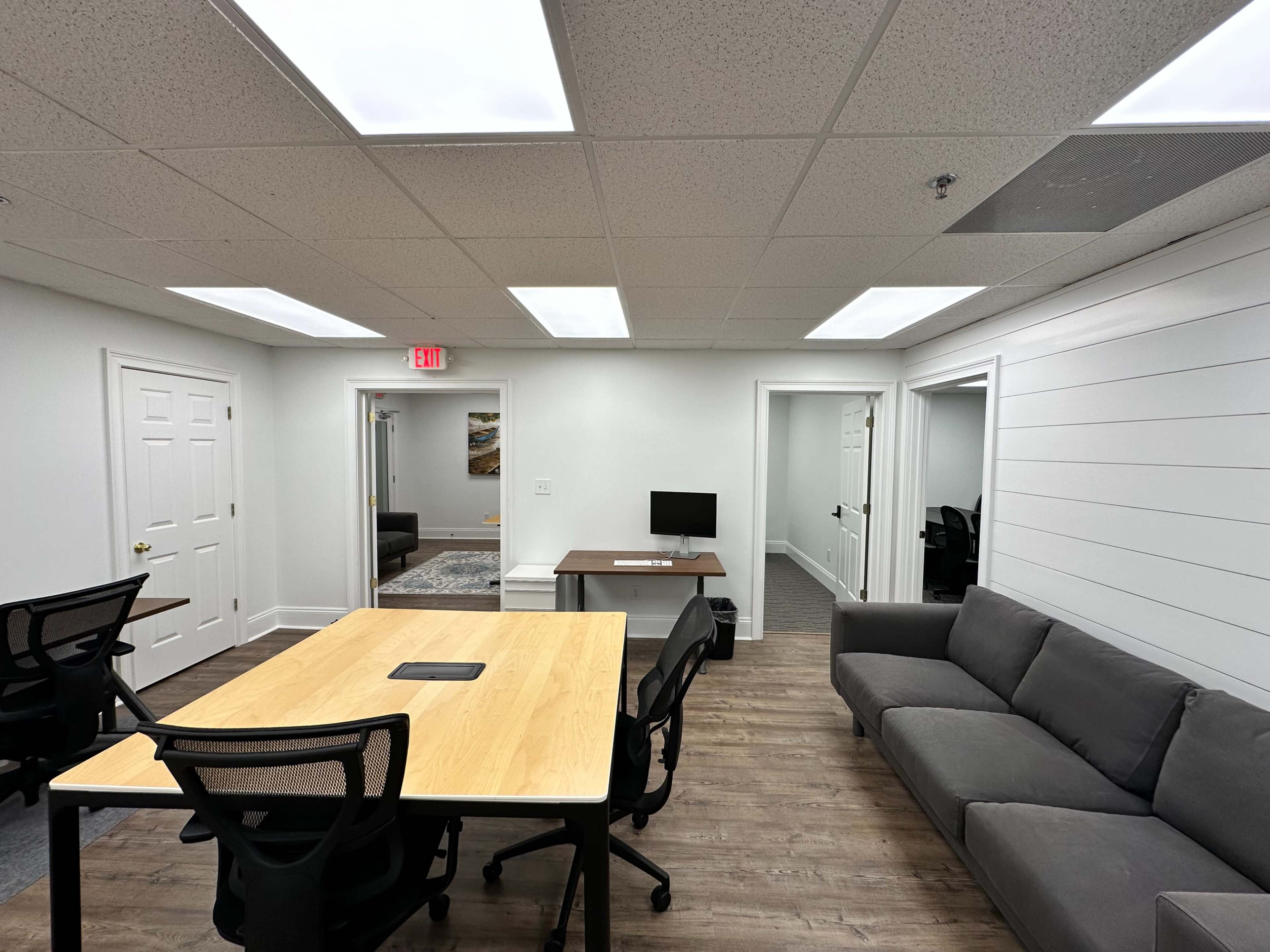 A modern office space with a wooden table, black chairs, a computer on a desk, and a gray sofa.