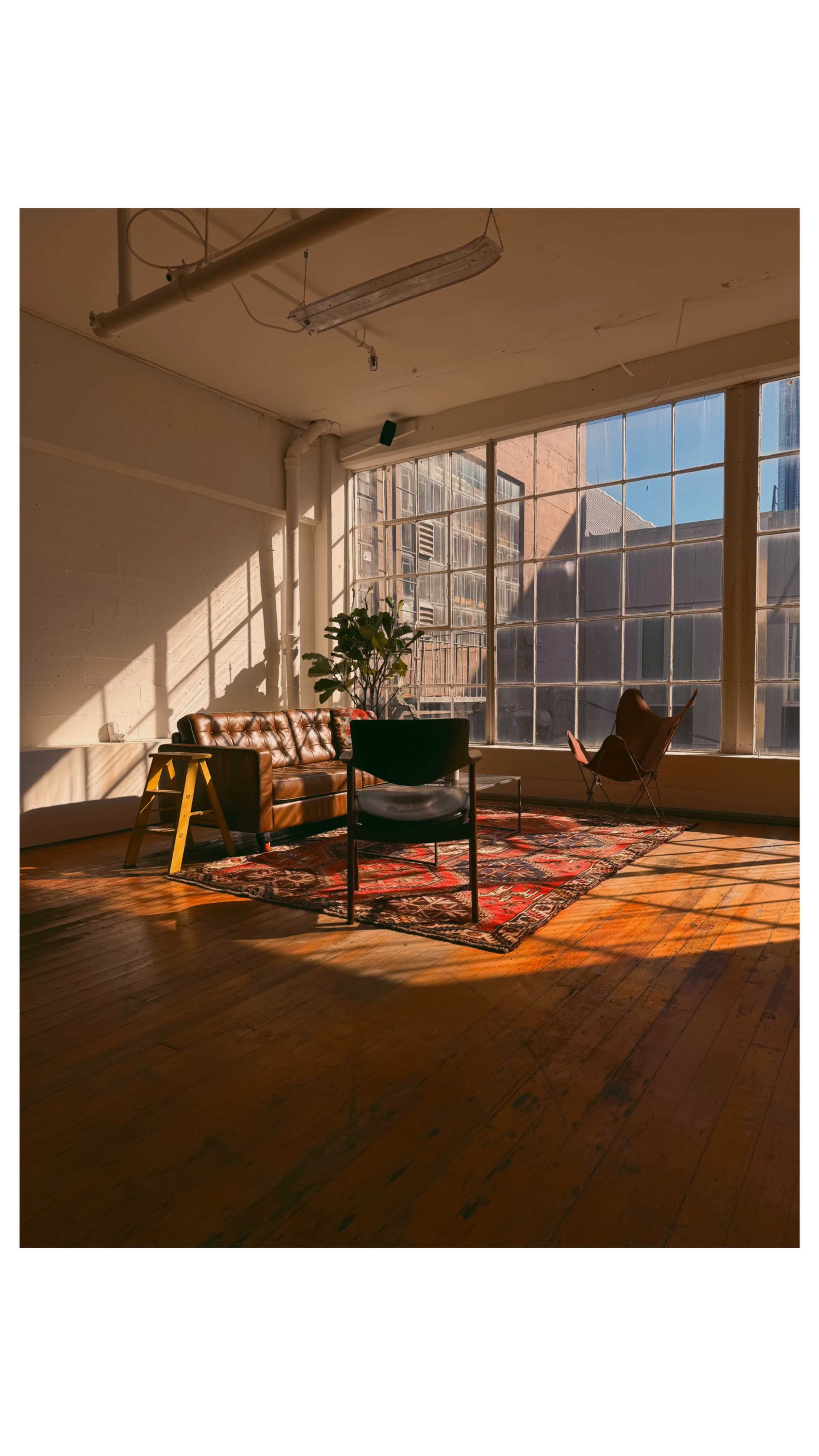 A cozy living space features a leather couch, armchair, and a large window that allows sunlight to cast shadows on the wooden floor.