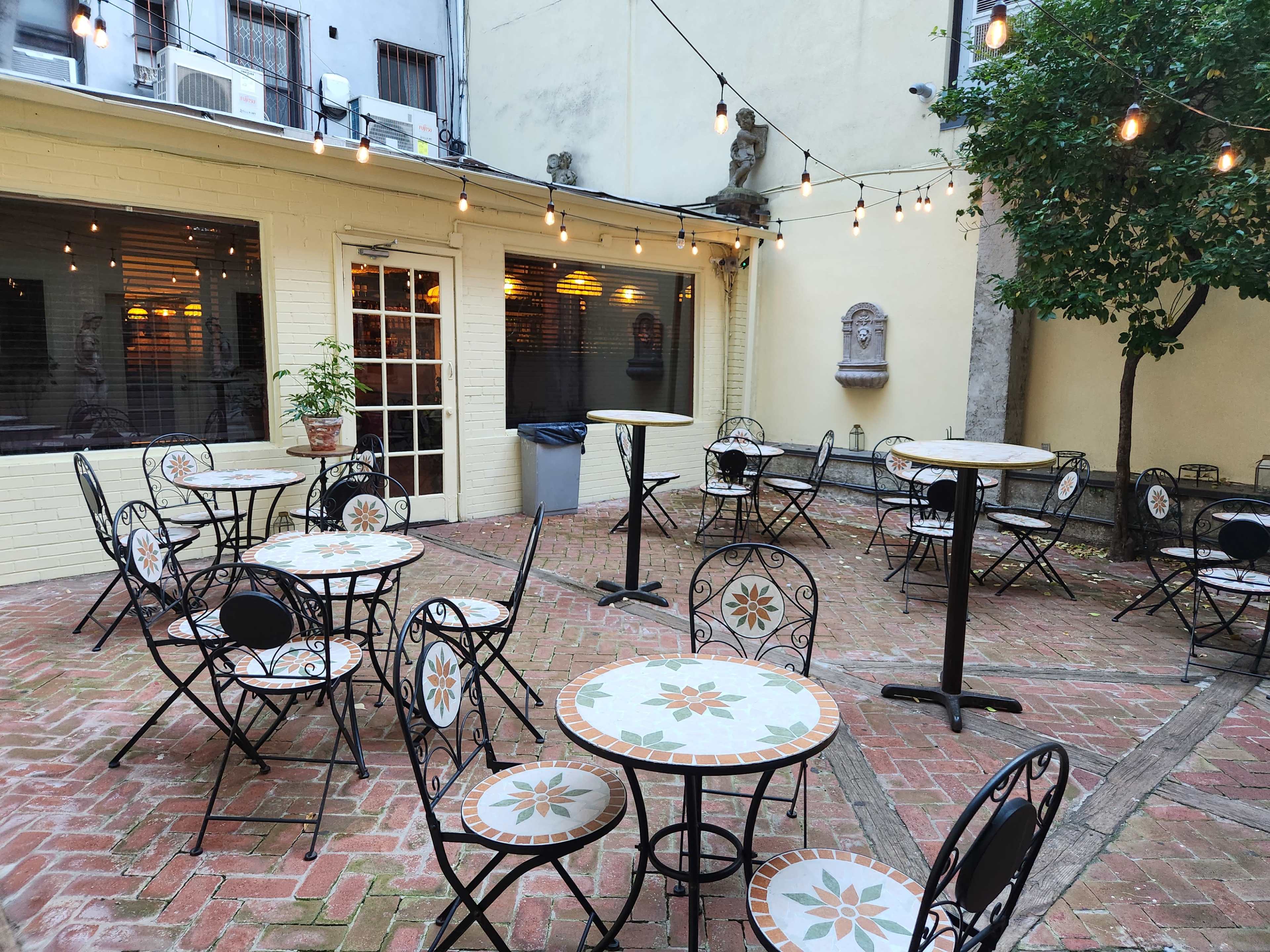 The image shows a courtyard with several black metal tables and chairs, illuminated by string lights, and features a brick floor with a small potted plant near the entrance.