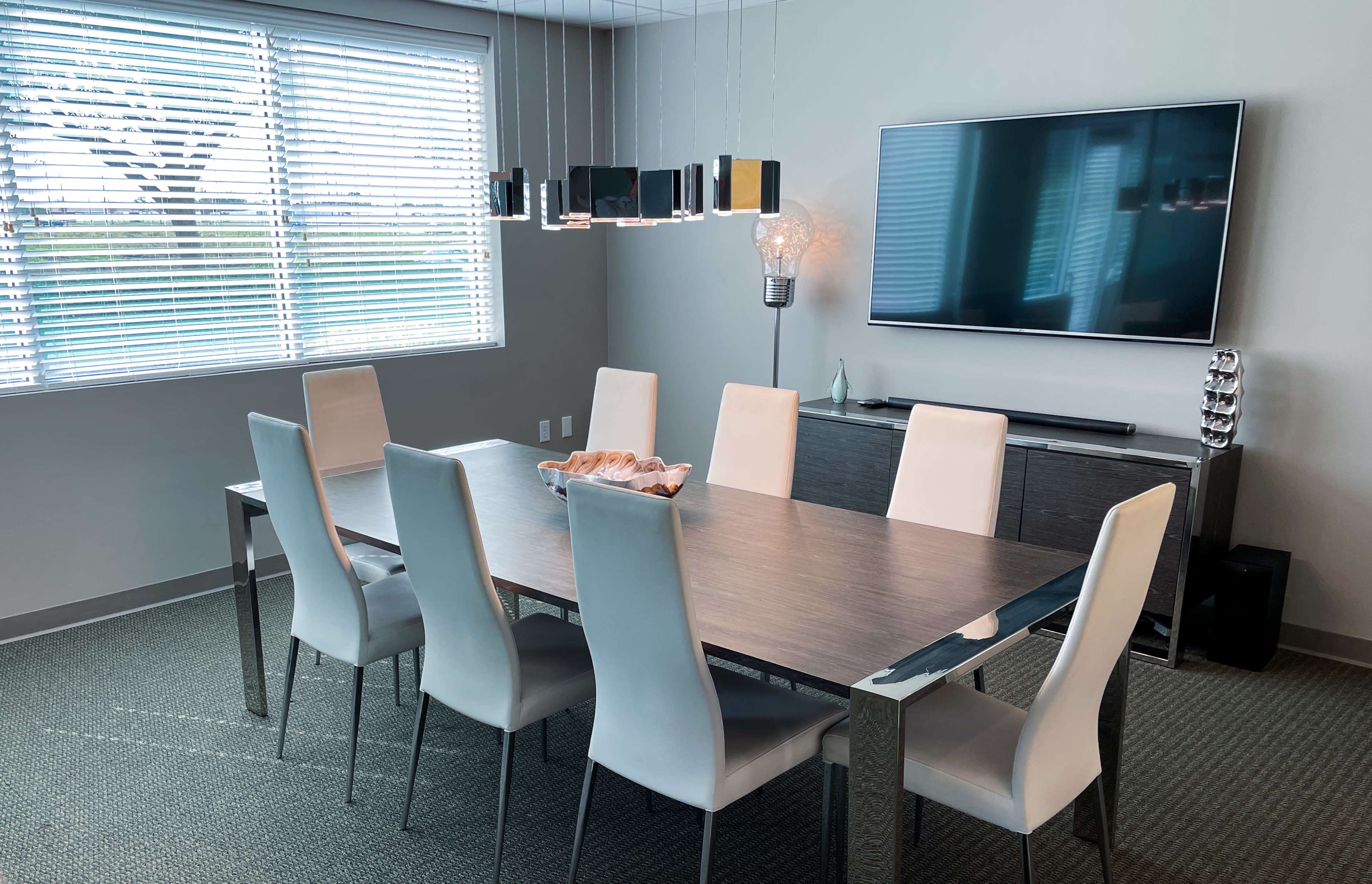 A modern dining room features a long table surrounded by eight light-colored chairs, with a television mounted on the wall and a window providing natural light.