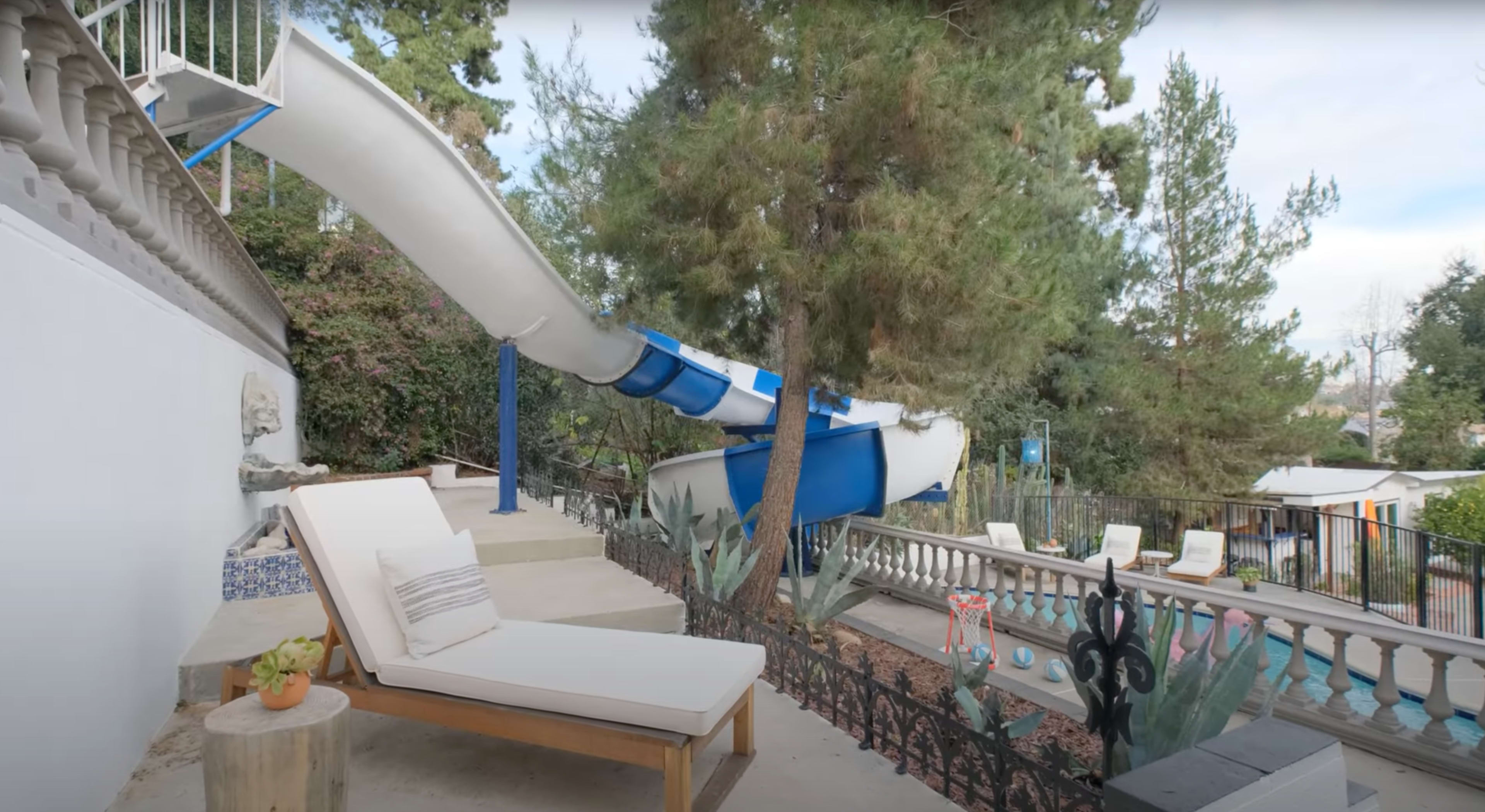 A curved blue water slide extends from a raised platform next to a sunbed, surrounded by pine trees and a fenced area with lounge chairs and a swimming pool in the background.