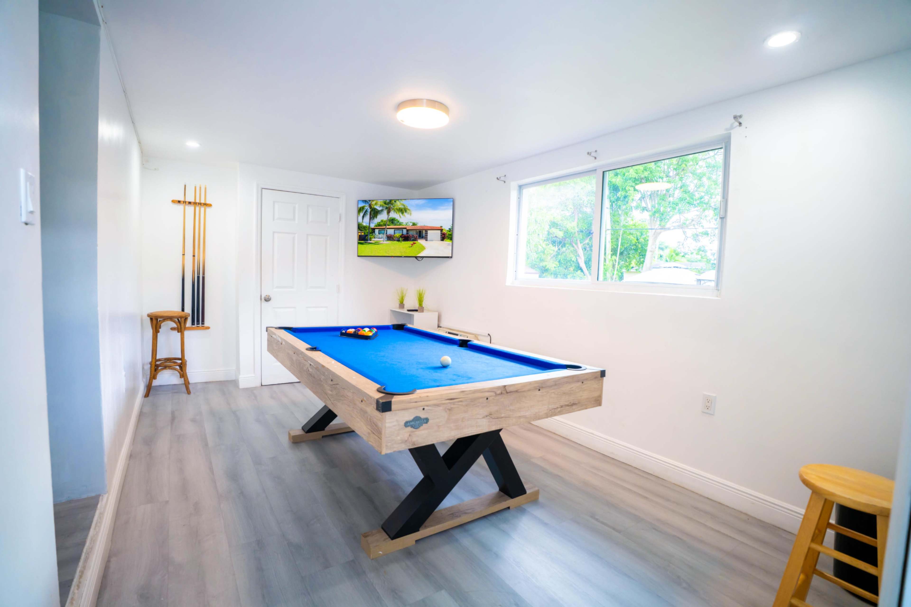 A bright game room features a blue pool table, a wall-mounted TV, and a door leading to another space.