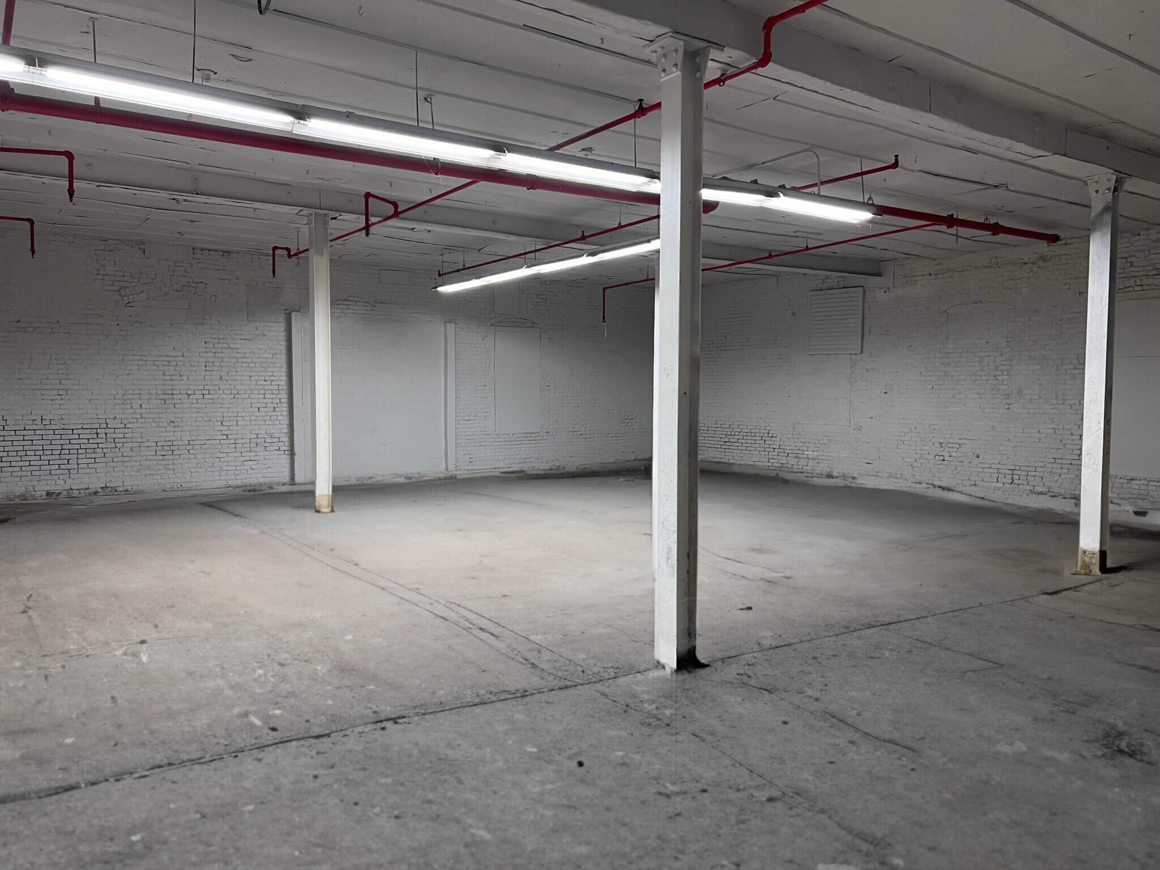 The image shows a vacant industrial space with exposed brick walls, concrete flooring, and fluorescent lighting.