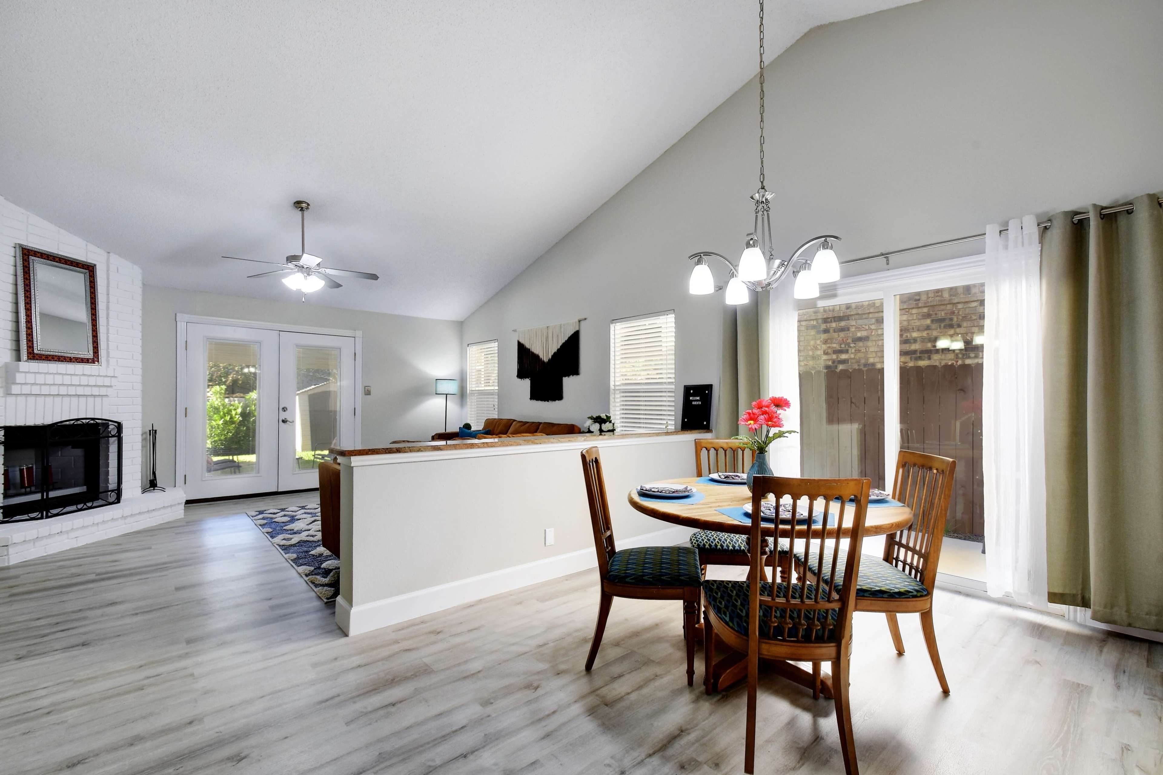 The image shows a modern living space featuring a dining table with chairs, a seating area, and a fireplace, all illuminated by natural light coming through sliding glass doors.