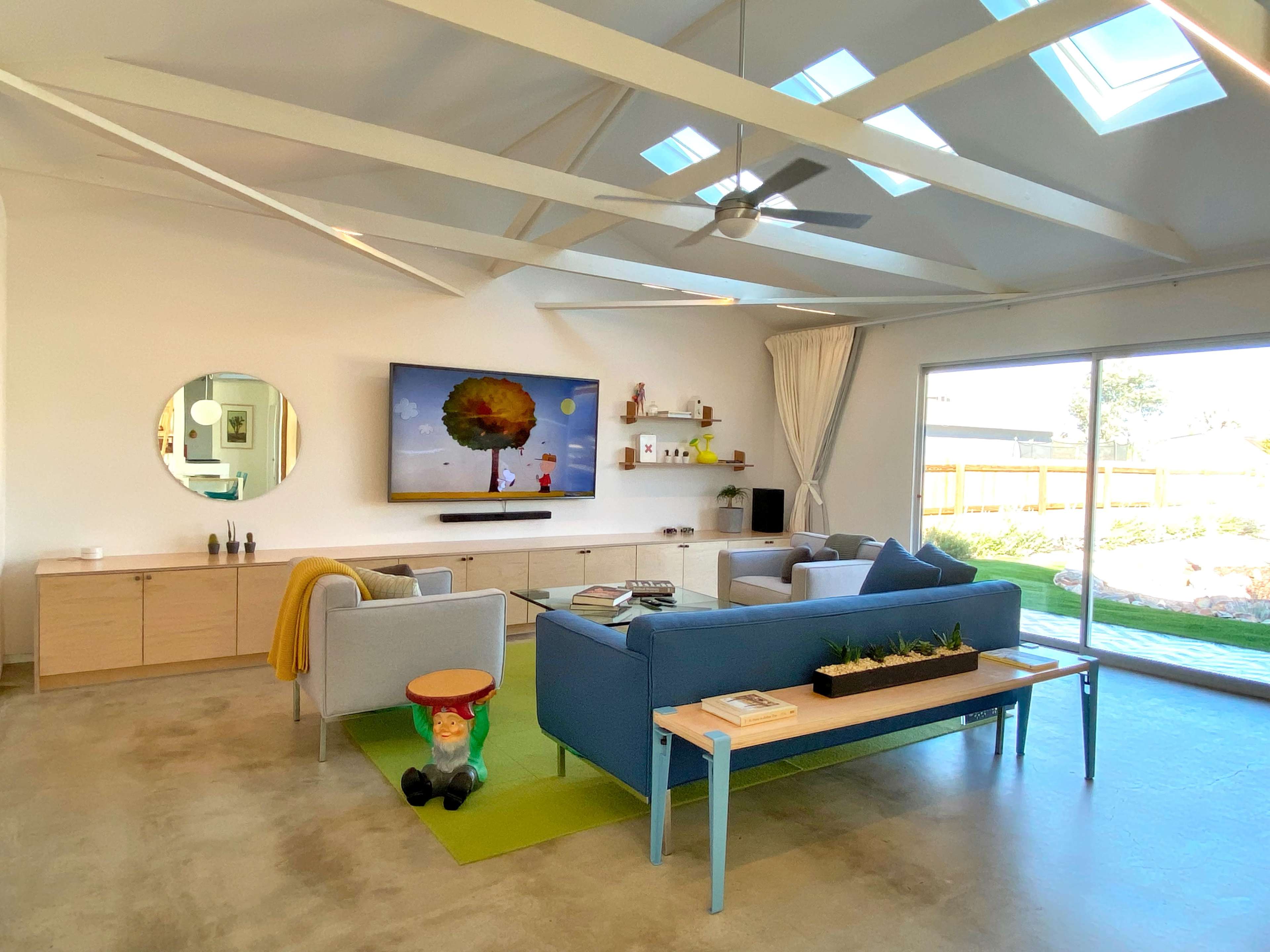 The image shows a modern living room featuring a blue sofa, a large television mounted on the wall, and a coffee table with a small planter, illuminated by natural light from skylights.