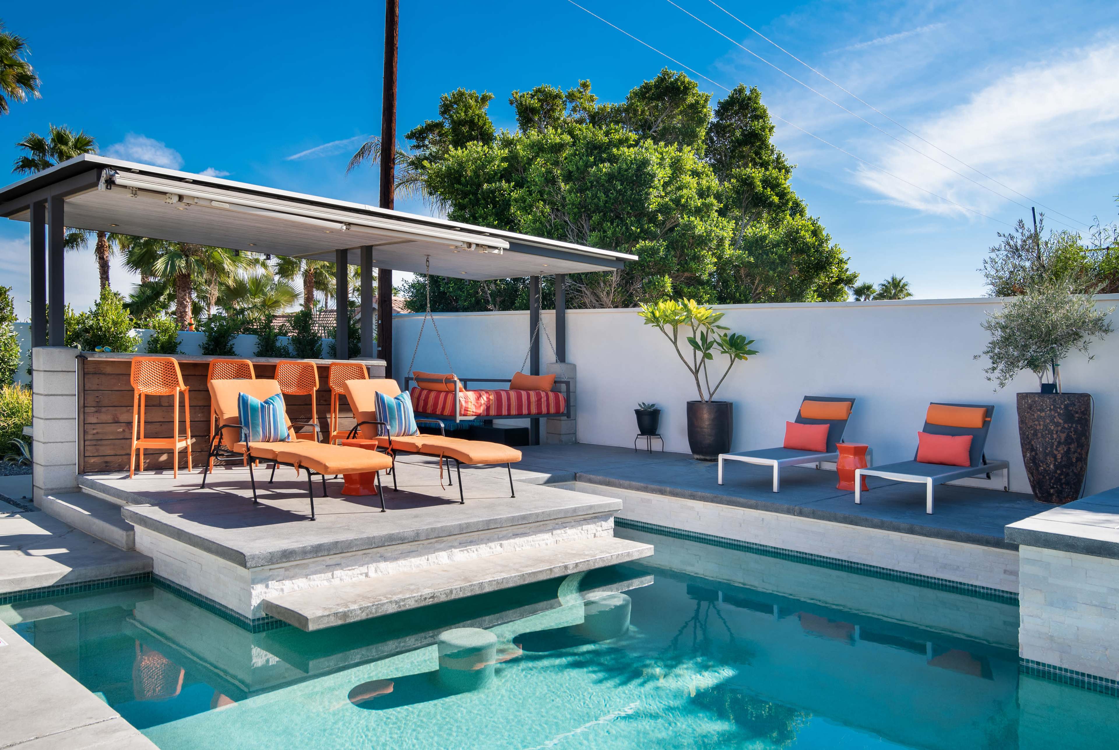 A modern pool area with lounge chairs, a covered seating space, and lush greenery under a clear blue sky.