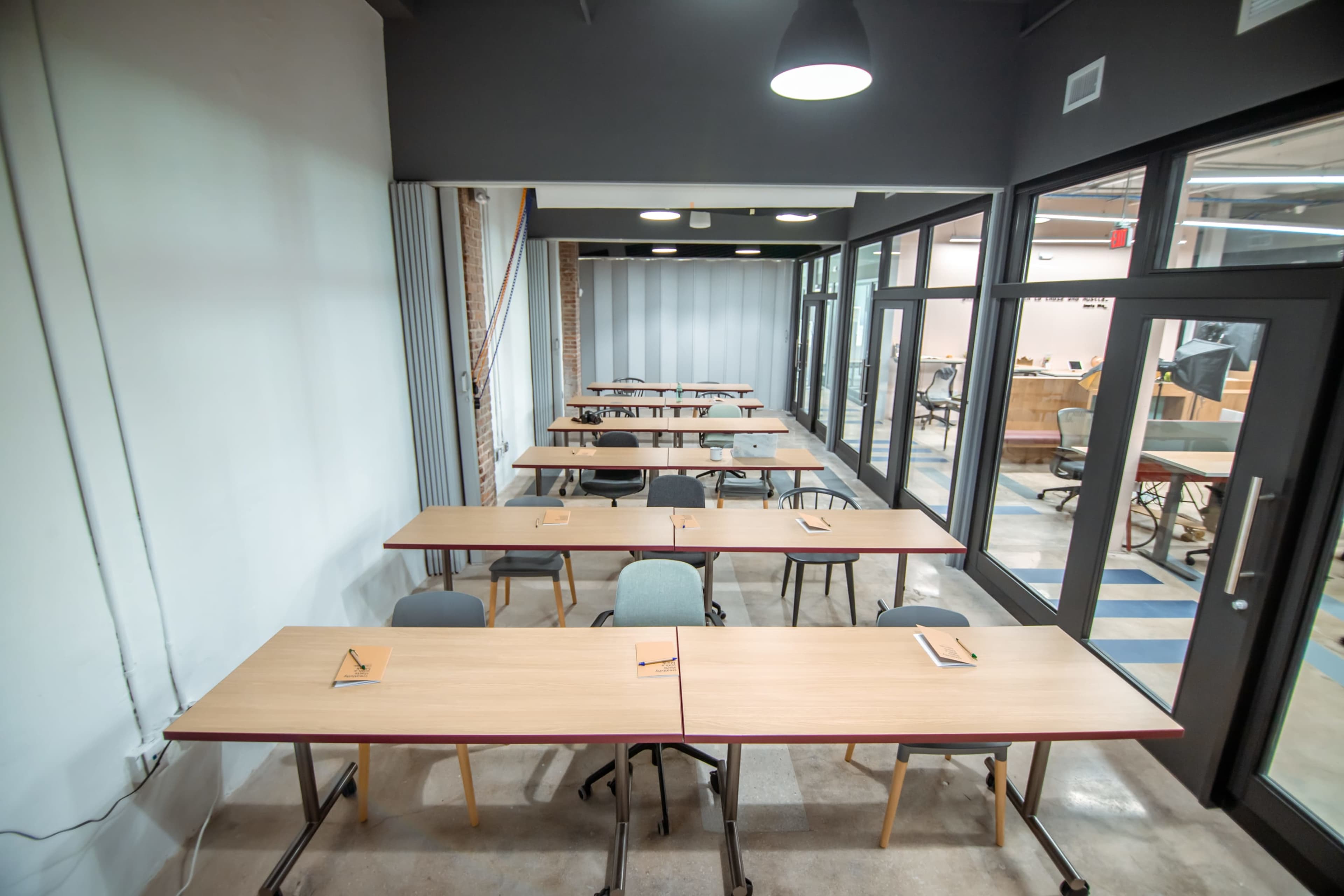 A spacious classroom with rectangular wooden tables arranged in rows, each set with a piece of paper and a pencil.