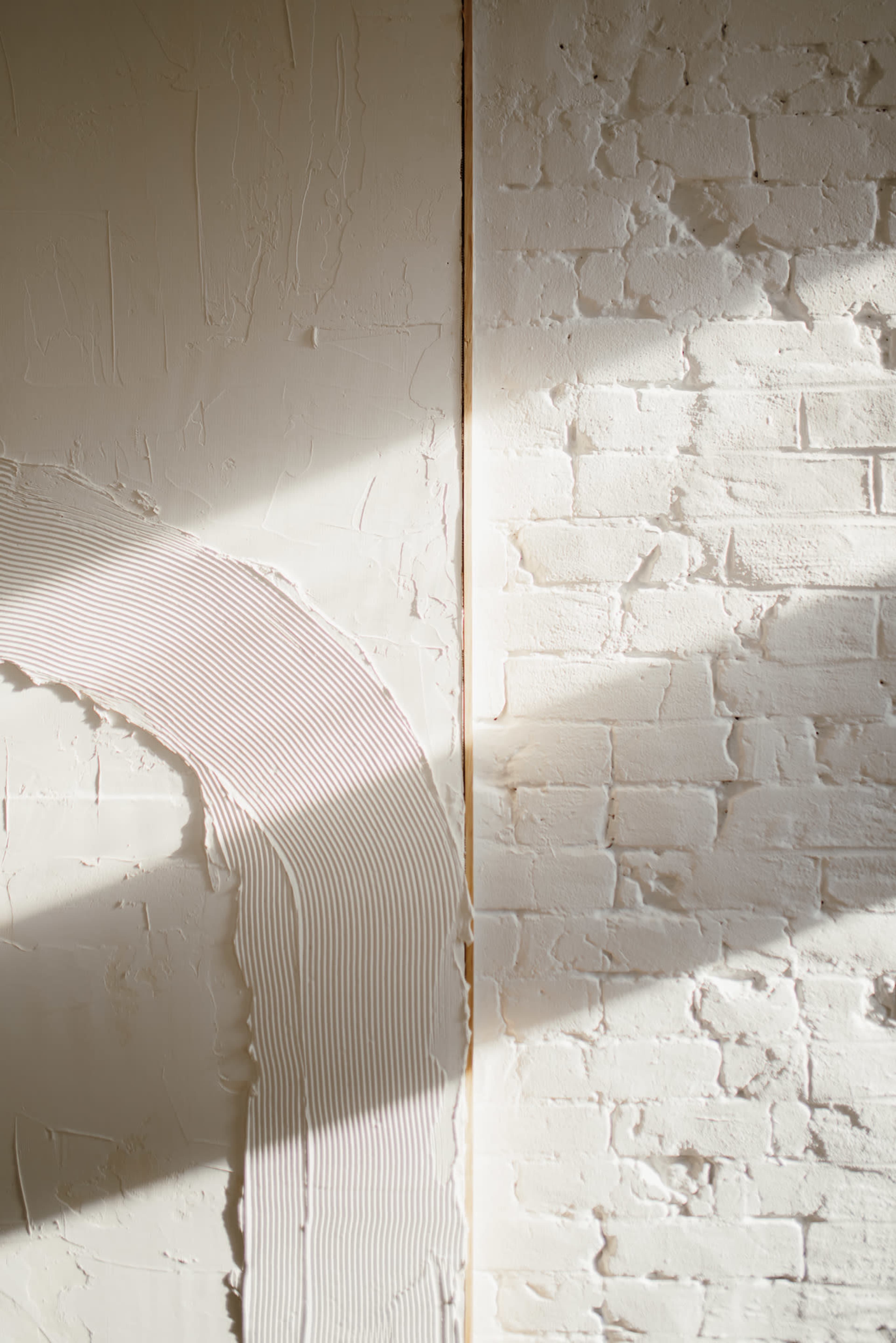 A textured wall featuring a curved application of plaster next to a brick surface illuminated by natural light.