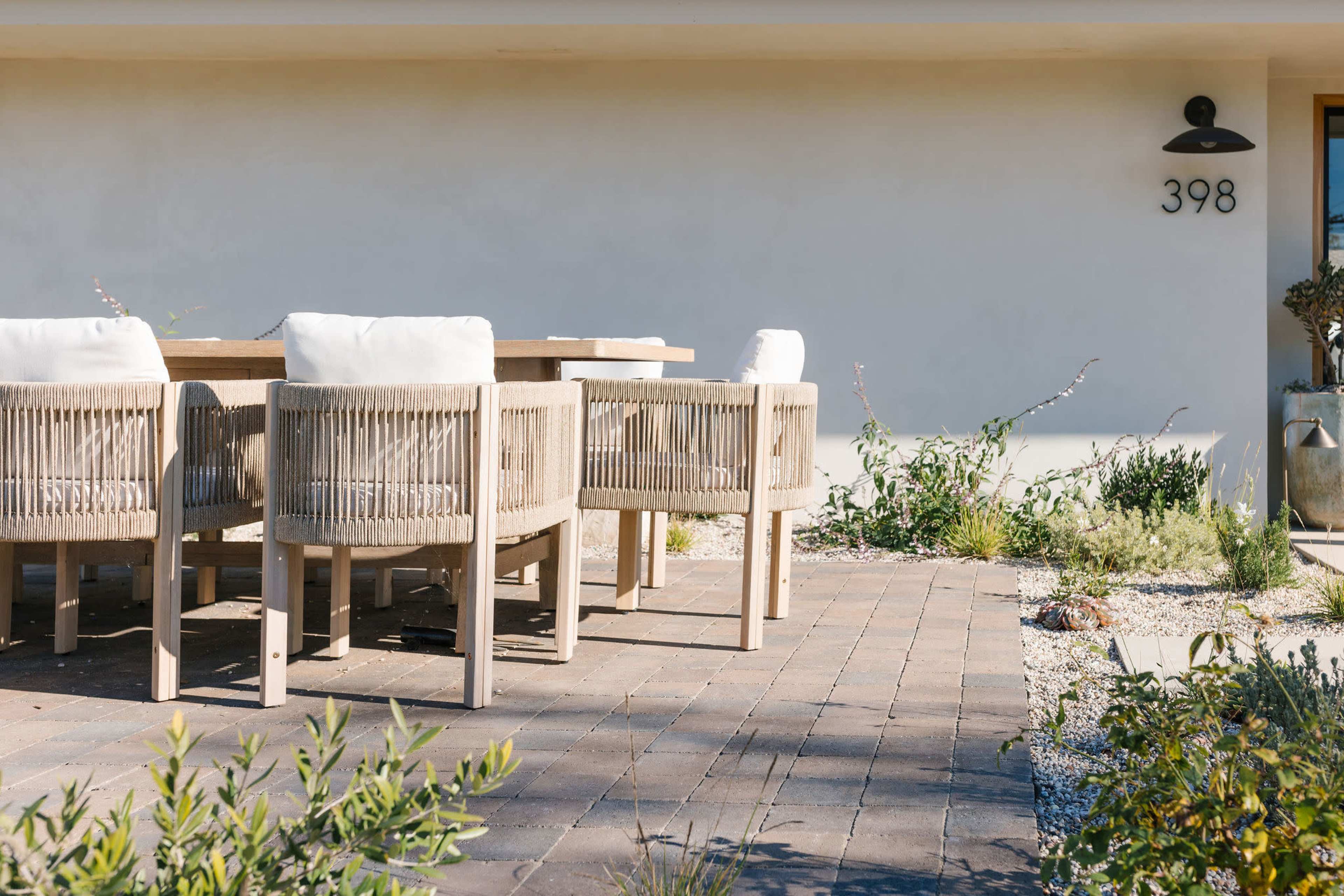 A modern outdoor dining area features a wooden table and six cushioned chairs on a paved patio beside plants and gravel.