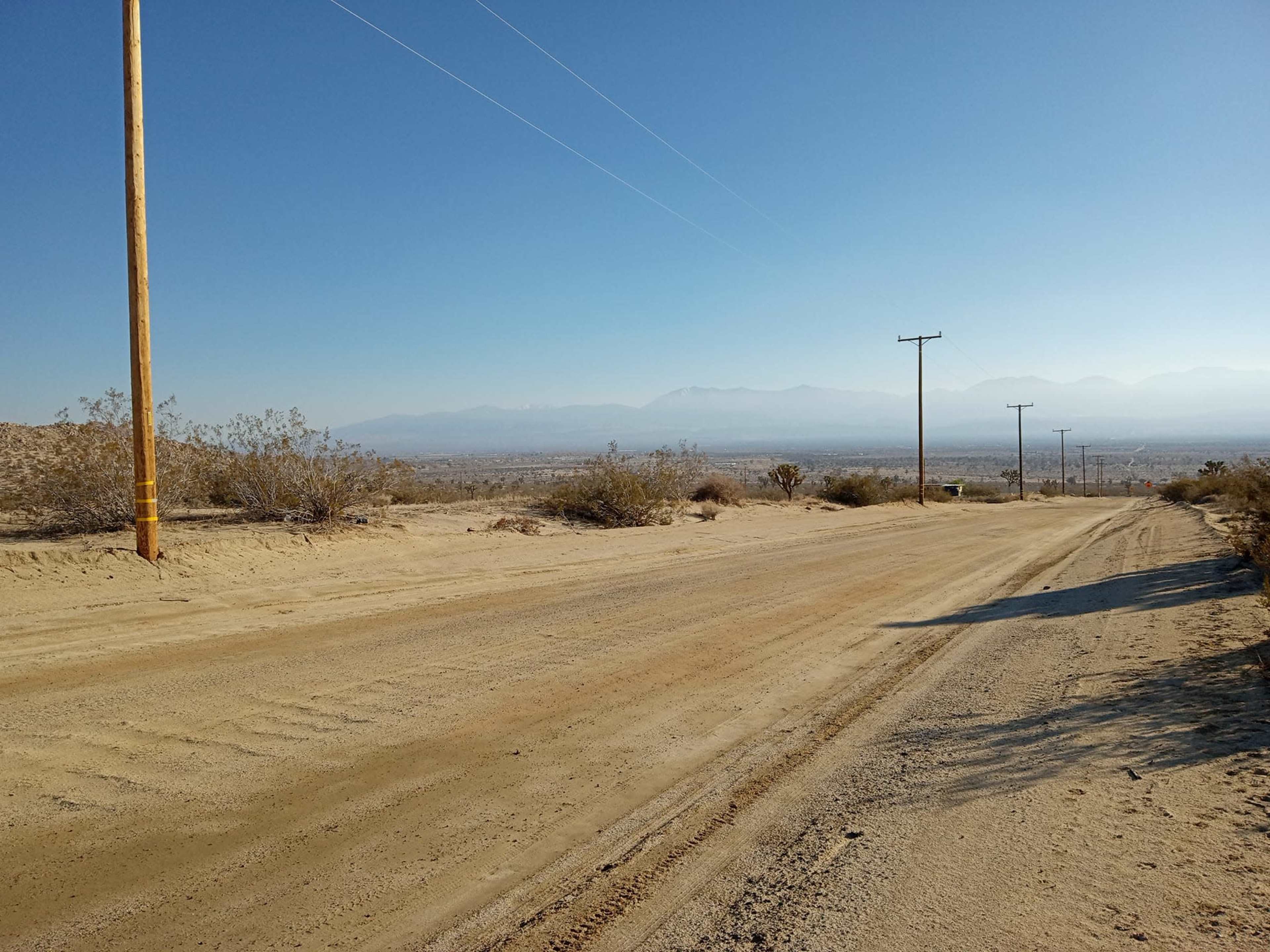 A dirt road stretches through a desert landscape, flanked by sparse vegetation and utility poles, with mountains visible in the distance.