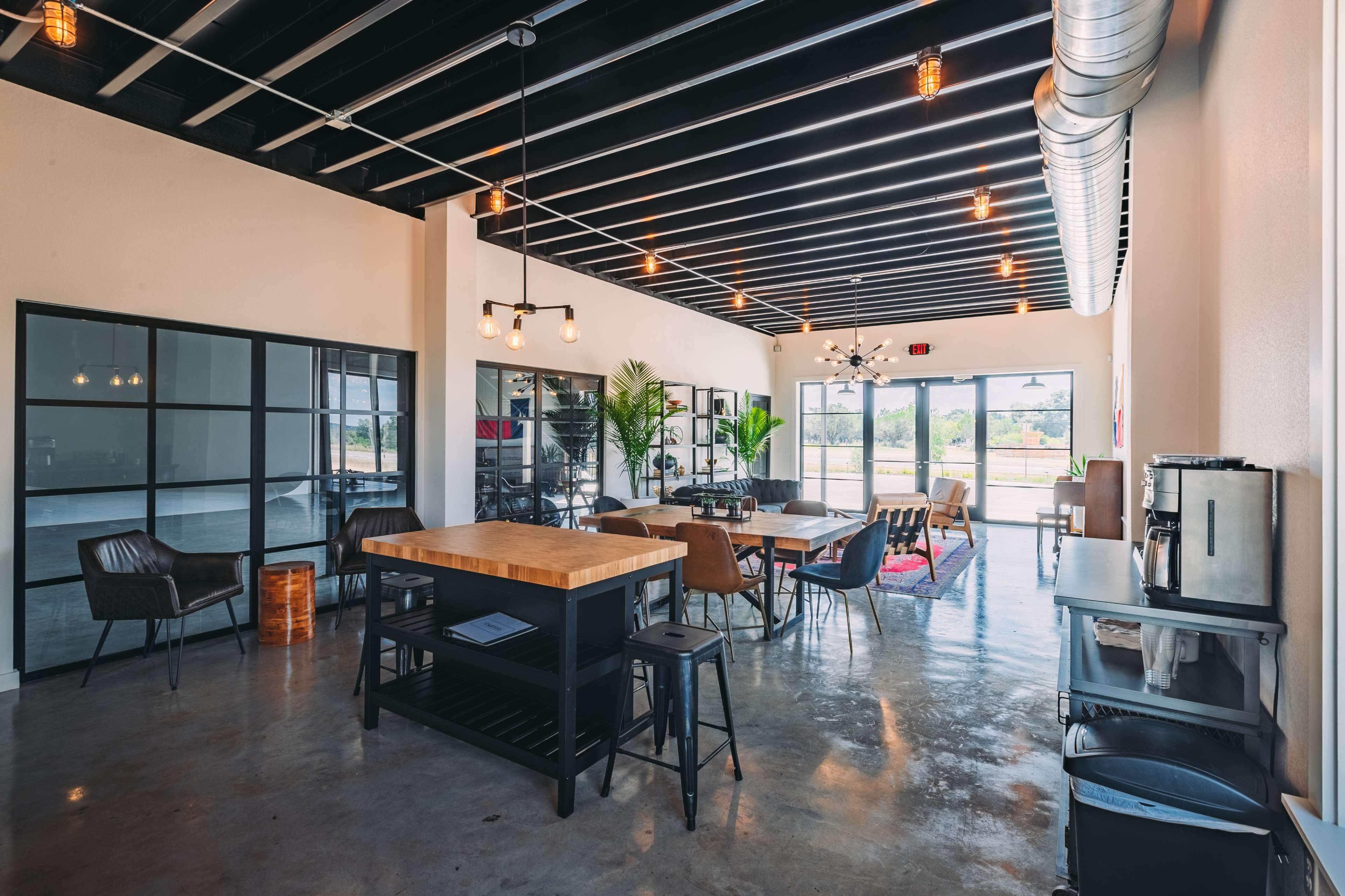 A modern office space features a mix of seating arrangements, a large wooden table, and large windows allowing natural light to illuminate the area.
