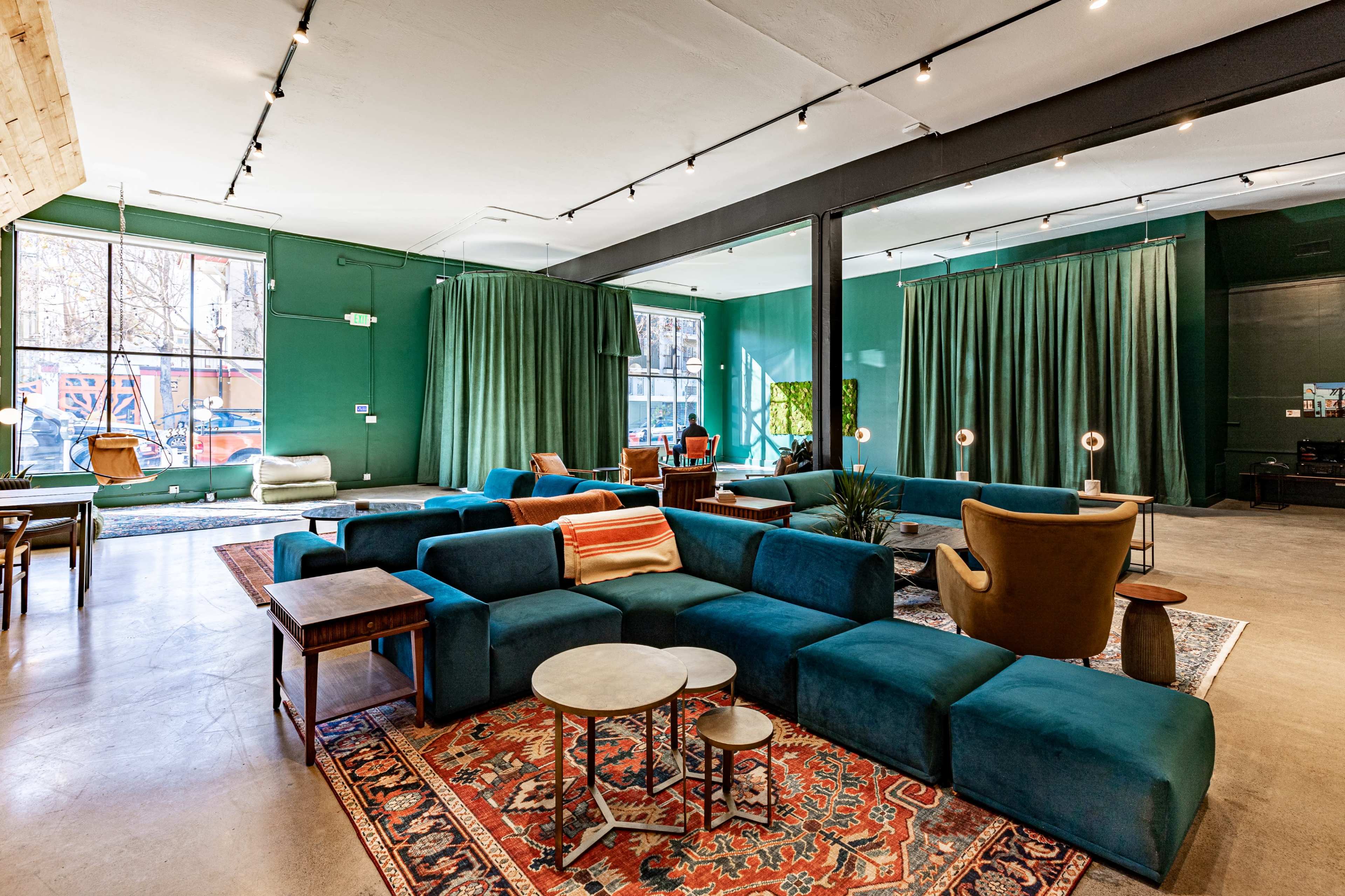 A spacious lounge area featuring green walls, a large blue sectional sofa, various seating options, and large windows with natural light.