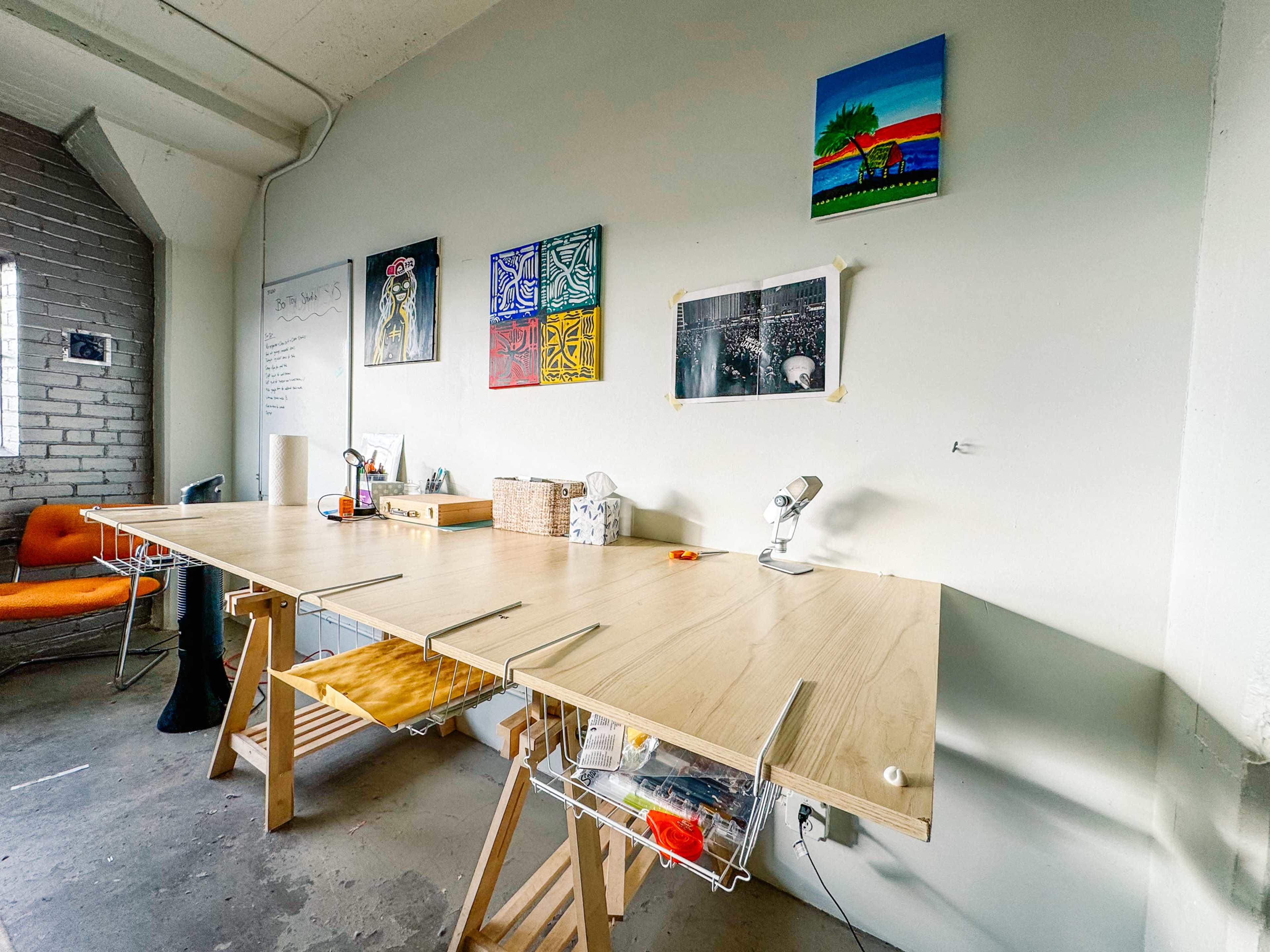 A wooden table with storage features is set against a light-colored wall, decorated with various artworks, in a bright workspace.