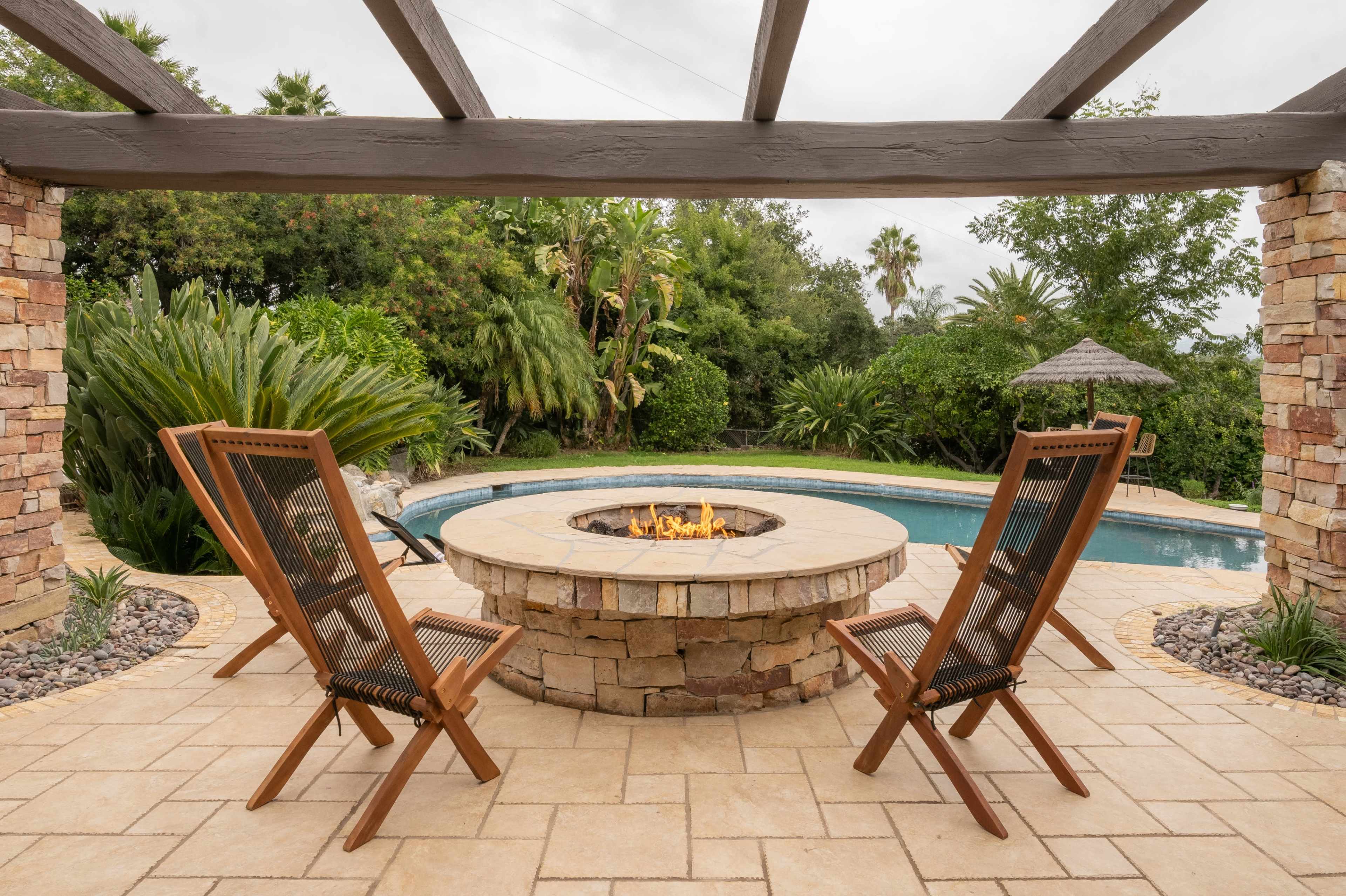 A stone fire pit surrounded by two wooden chairs, overlooking a pool and lush greenery in a residential backyard.