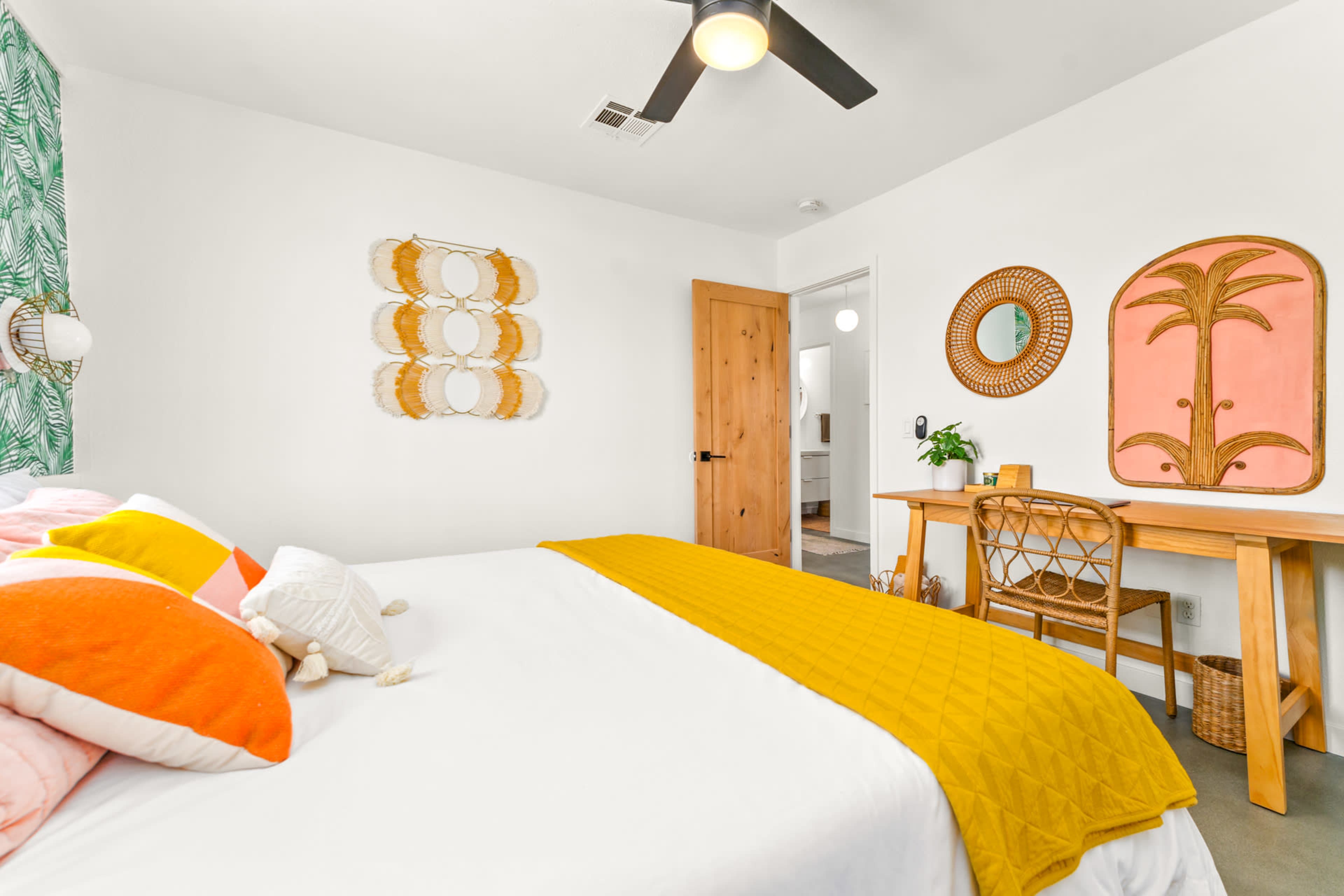 A bright bedroom features a bed with colorful pillows, a wooden desk, and decor of leafy patterns on the wall.