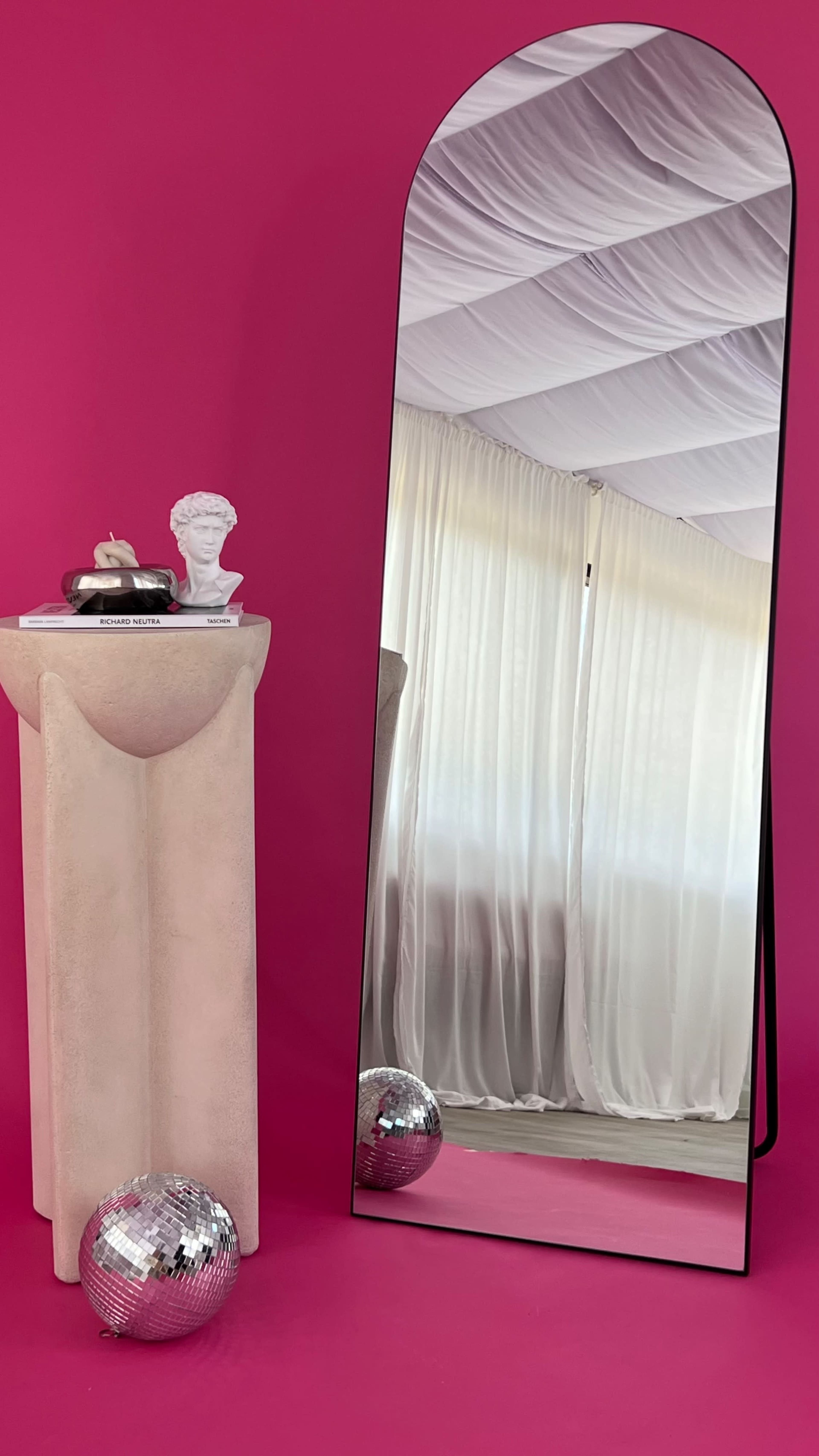 The image features a tall, arched mirror leaning against a bright pink wall, next to a sculptural pedestal with decor items and a disco ball on the floor.