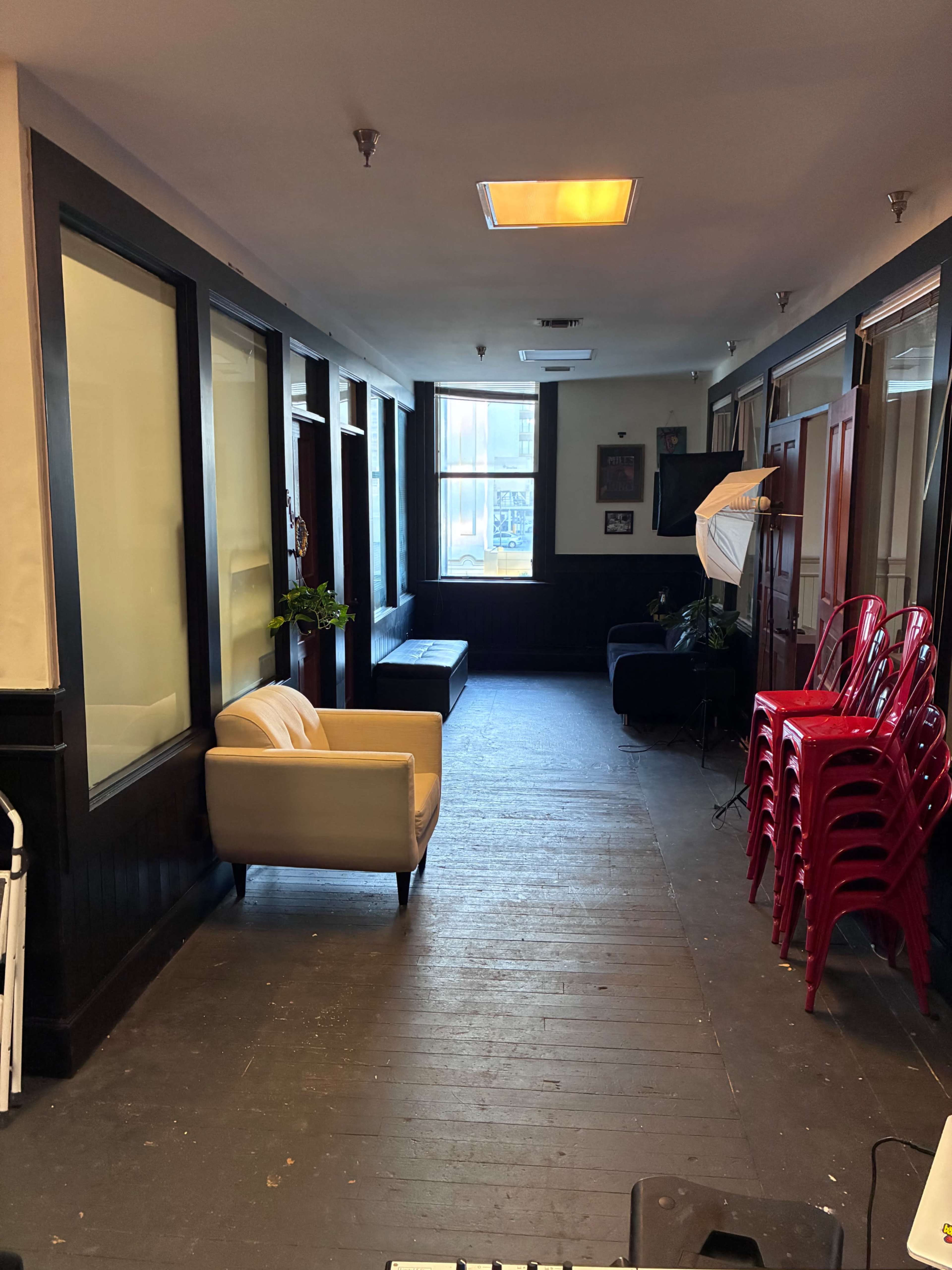 The image shows a narrow hallway with seating areas on either side, including a couch and stacked red chairs against the wall.