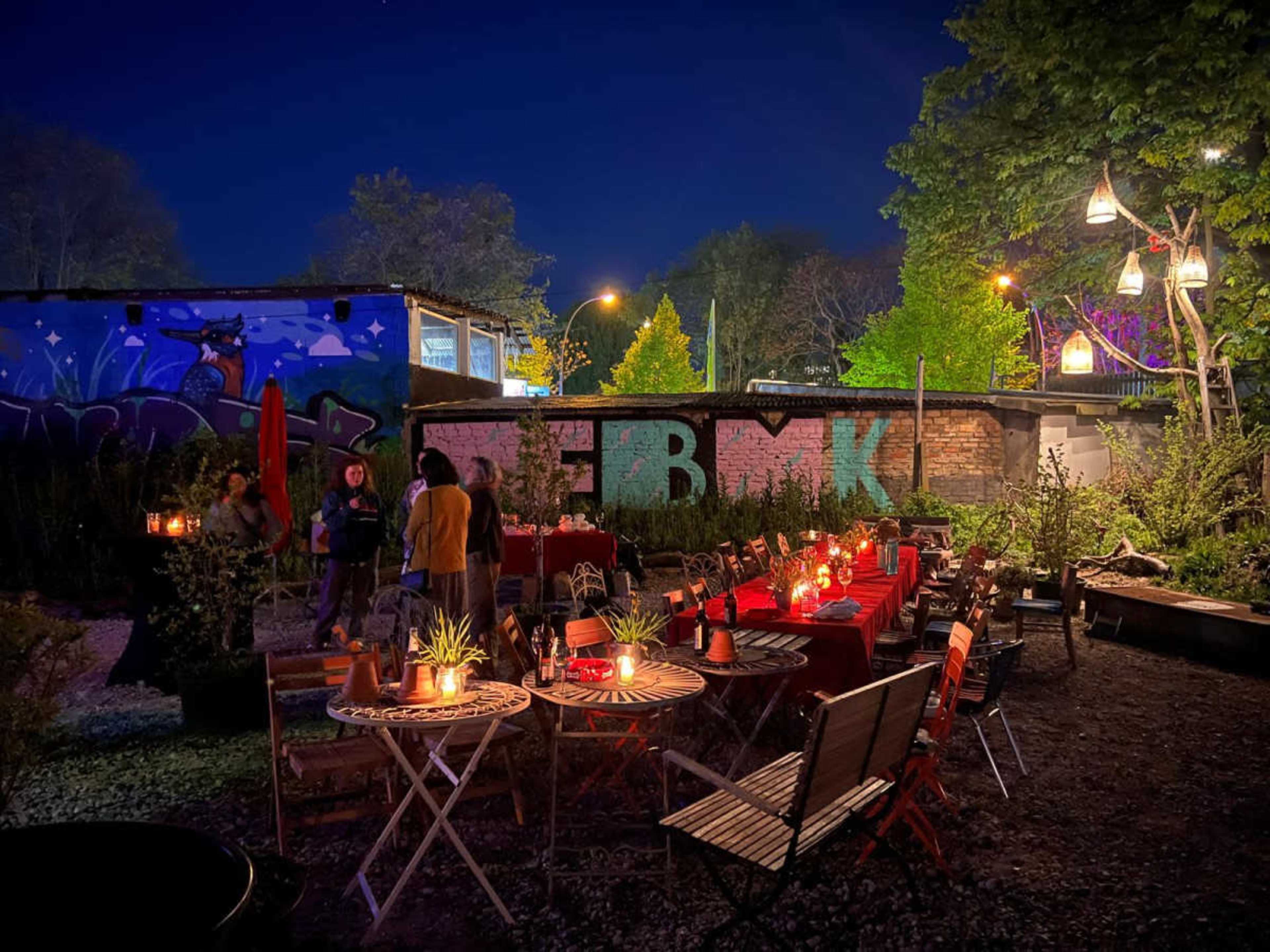 A dimly lit outdoor venue features tables with candles surrounded by greenery and colorful graffiti in the background.