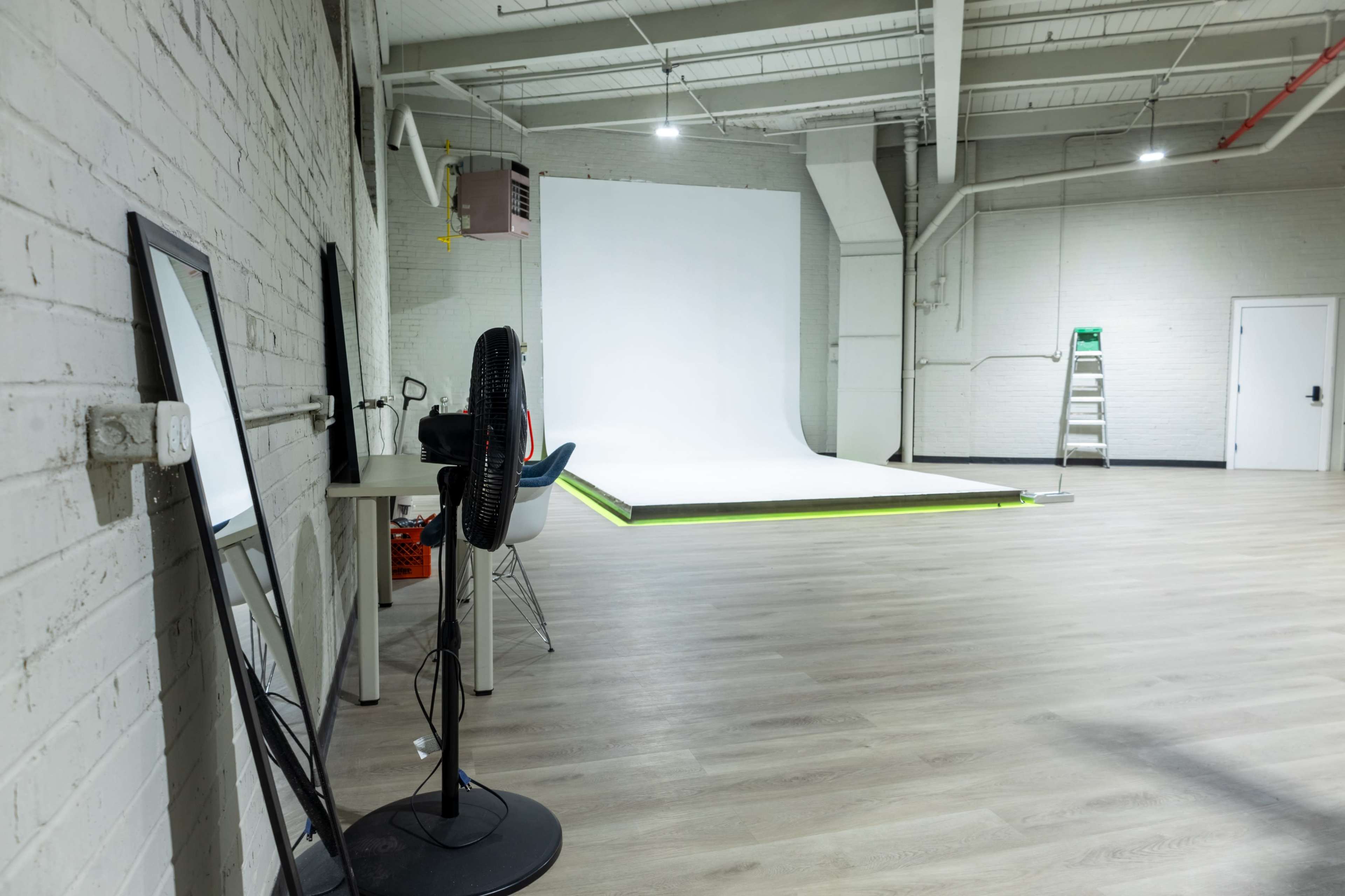 The image shows a spacious photography studio featuring a white backdrop, a fan, a mirror, and a ladder against the wall.